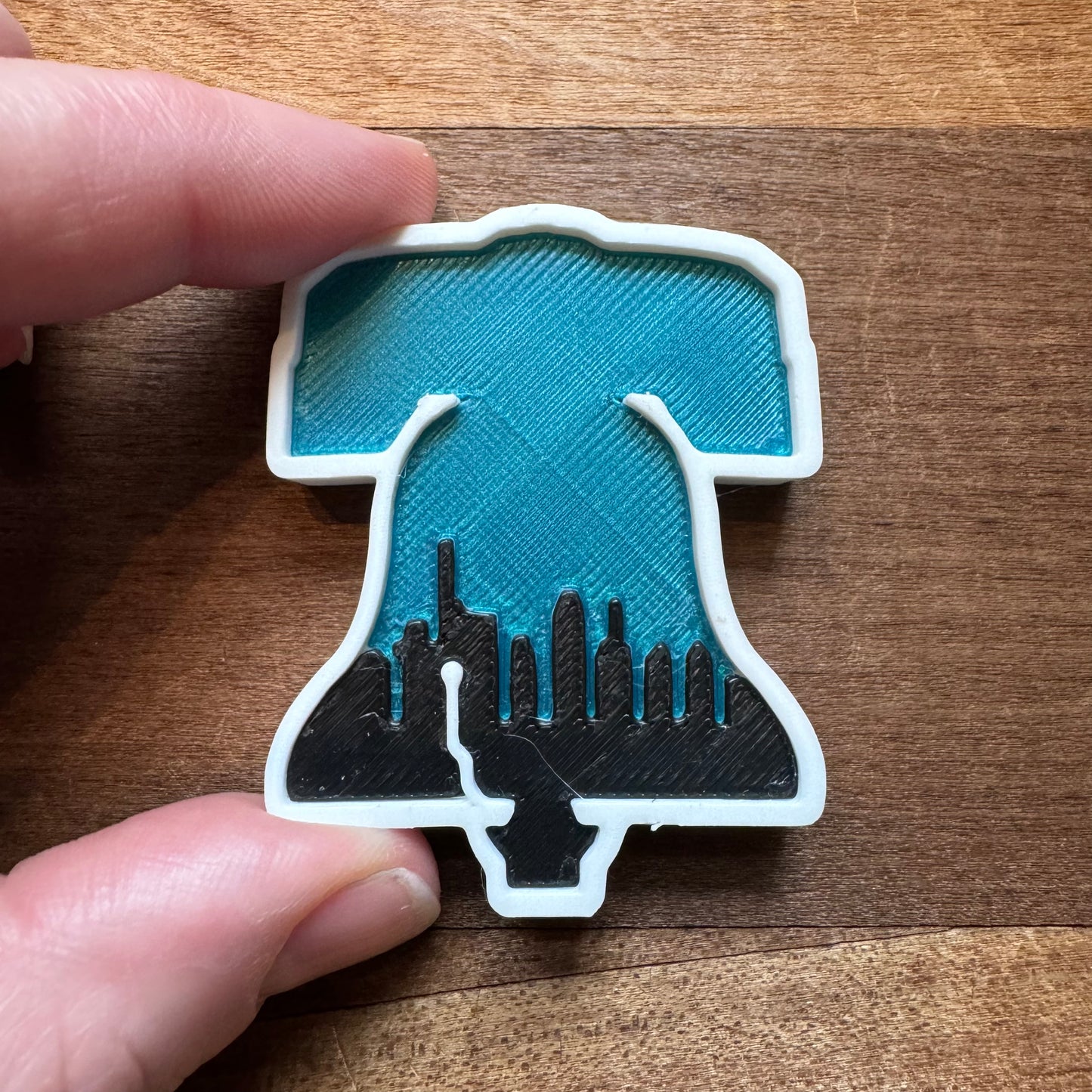 Close-up of a hand holding a small 3D-printed "Philly Magnets" by Rosewood Home, shaped like the Liberty Bell, featuring a blue interior and a black Philadelphia Skyline at the bottom.