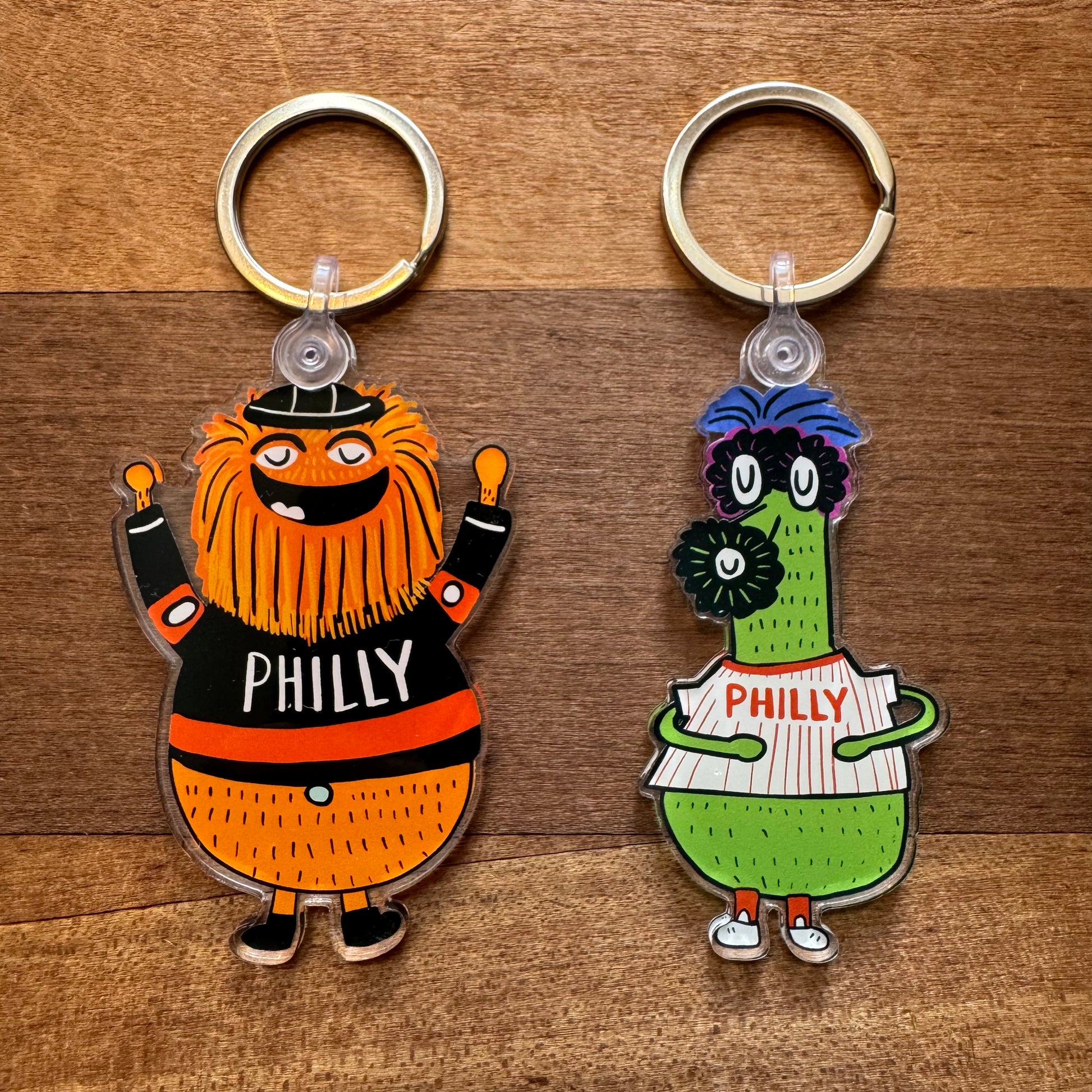 The Gritty & Phanatic Keychains from Ana Thorne feature two vibrant characters: an orange, bearded figure in a black "Philly" shirt and a green, furry character with glasses in a white "Philly" jersey.