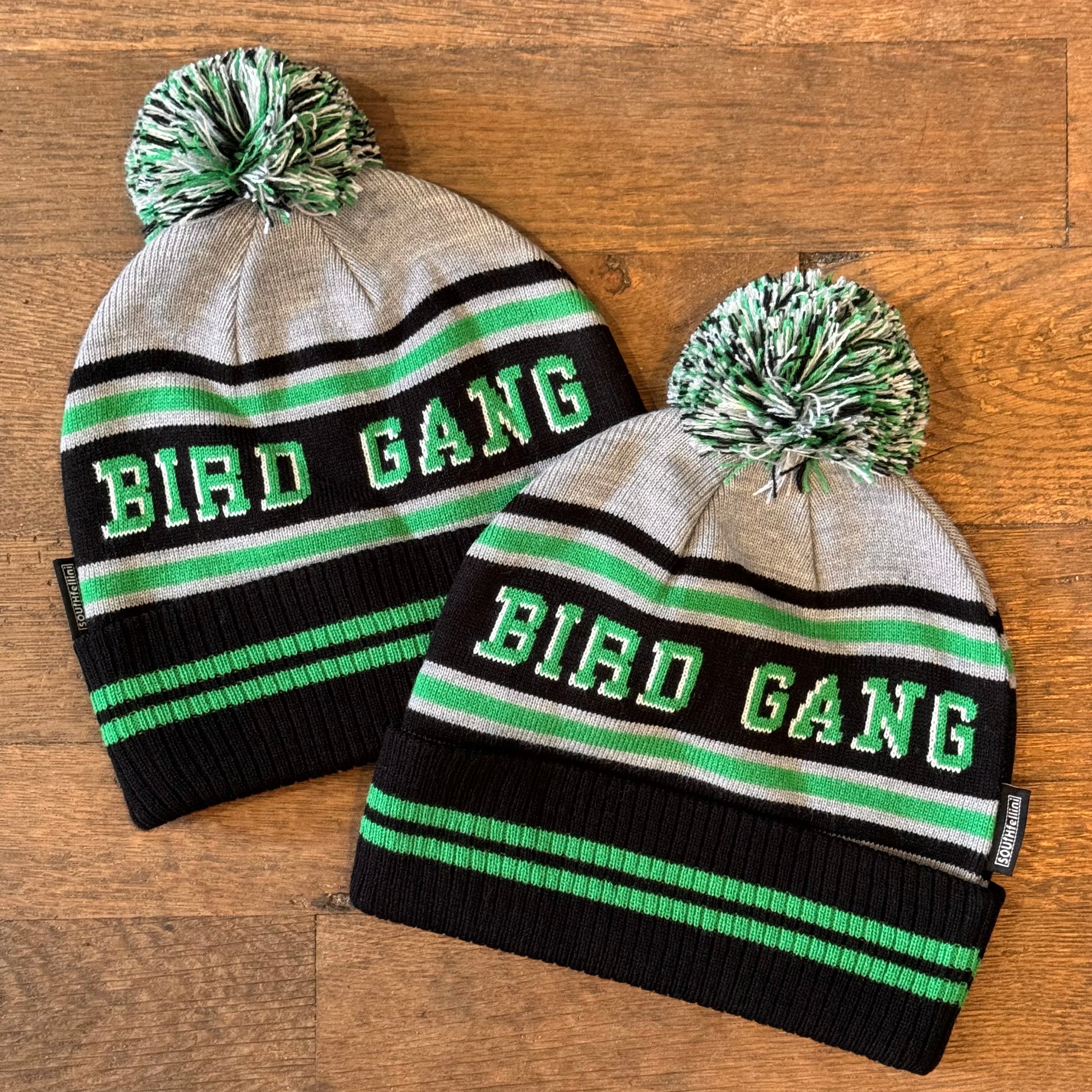 Two Bird Gang Eagles Beanies by South Fellini are perfect for any Eagles fan. Both hats feature gray, black, green, and white stripes with the text "BIRD GANG" on the front. These knit beanies are beautifully displayed on a wooden surface.