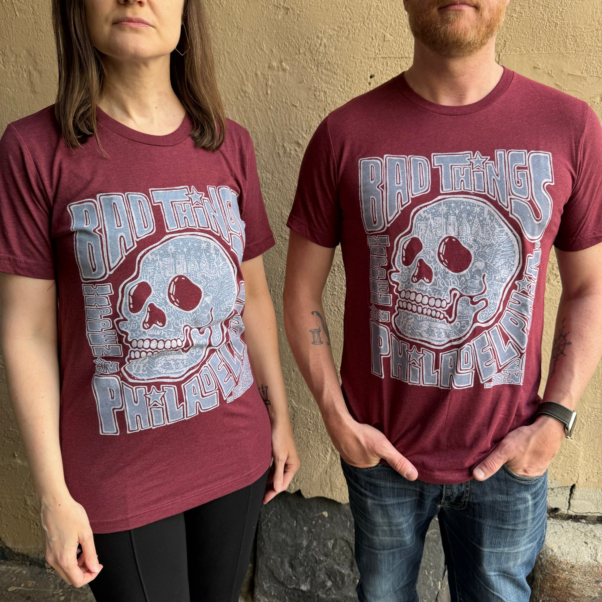 Two people wearing super soft unisex matching maroon Bad Things Happen T-Shirts by Paul Carpenter with a skull design and the words "BAD THINGS" in bold letters against a textured beige wall.