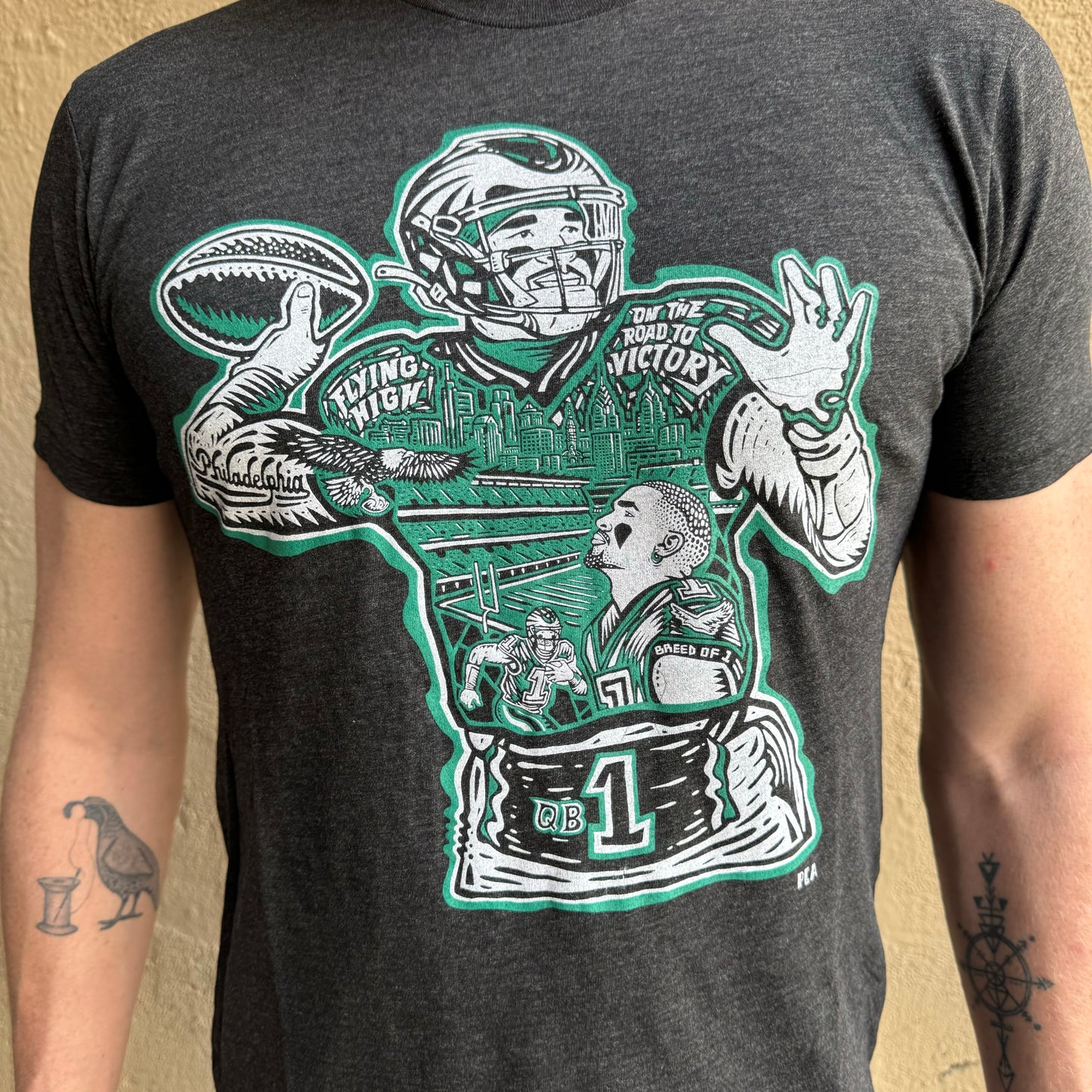 A person wearing a dark grey Jalen Hurts T-Shirt from Paul Carpenter, showcasing an illustrated design featuring a football player, cityscape skyline design, and various football-related elements in green and white.