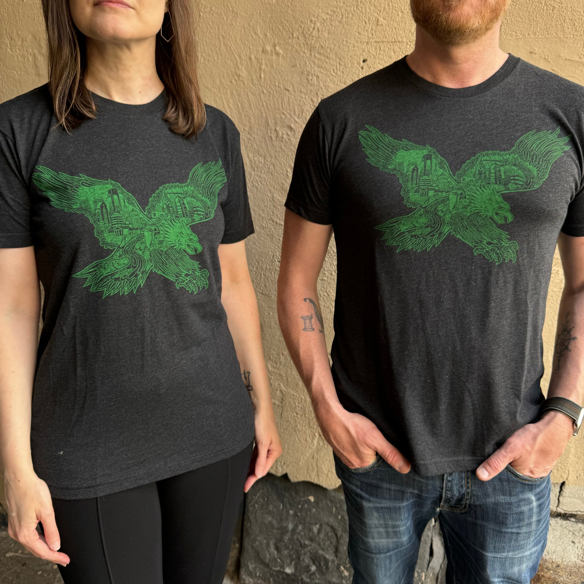 Two people standing side by side, both wearing black Swooping Eagle T-Shirts with a green bird graphic from Paul Carpenter. The person on the left has long hair; the person on the right has a beard and tattoos on their forearm, perhaps showcasing Paul Carpenter Art.