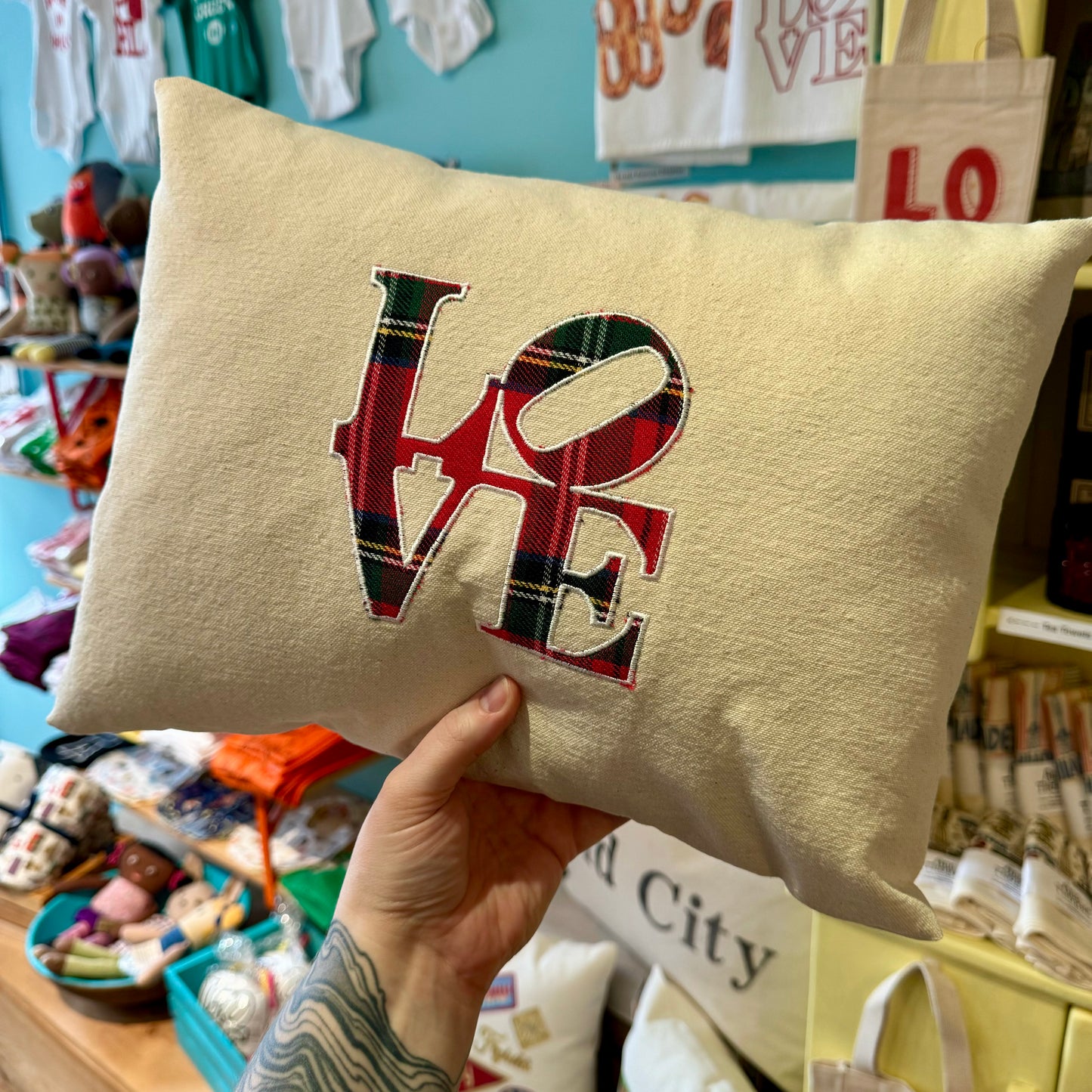 Person holding a beige Holiday Plaid Pillow by The Pillow Works, featuring the word "LOVE" in vibrant plaid letters, in a store with various festive merchandise in the background.