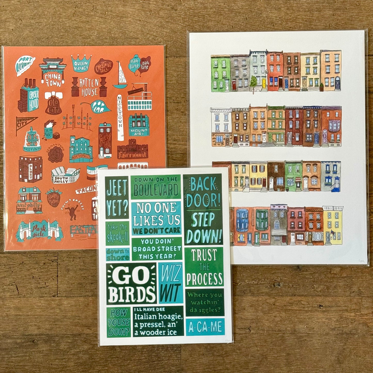 A set of three Philadelphia Prints from Kate Otte: one featuring Philly-themed stickers, another depicting colorful row houses, and a third highlighting various Philly lingo and phrases, all displayed on a wooden surface.