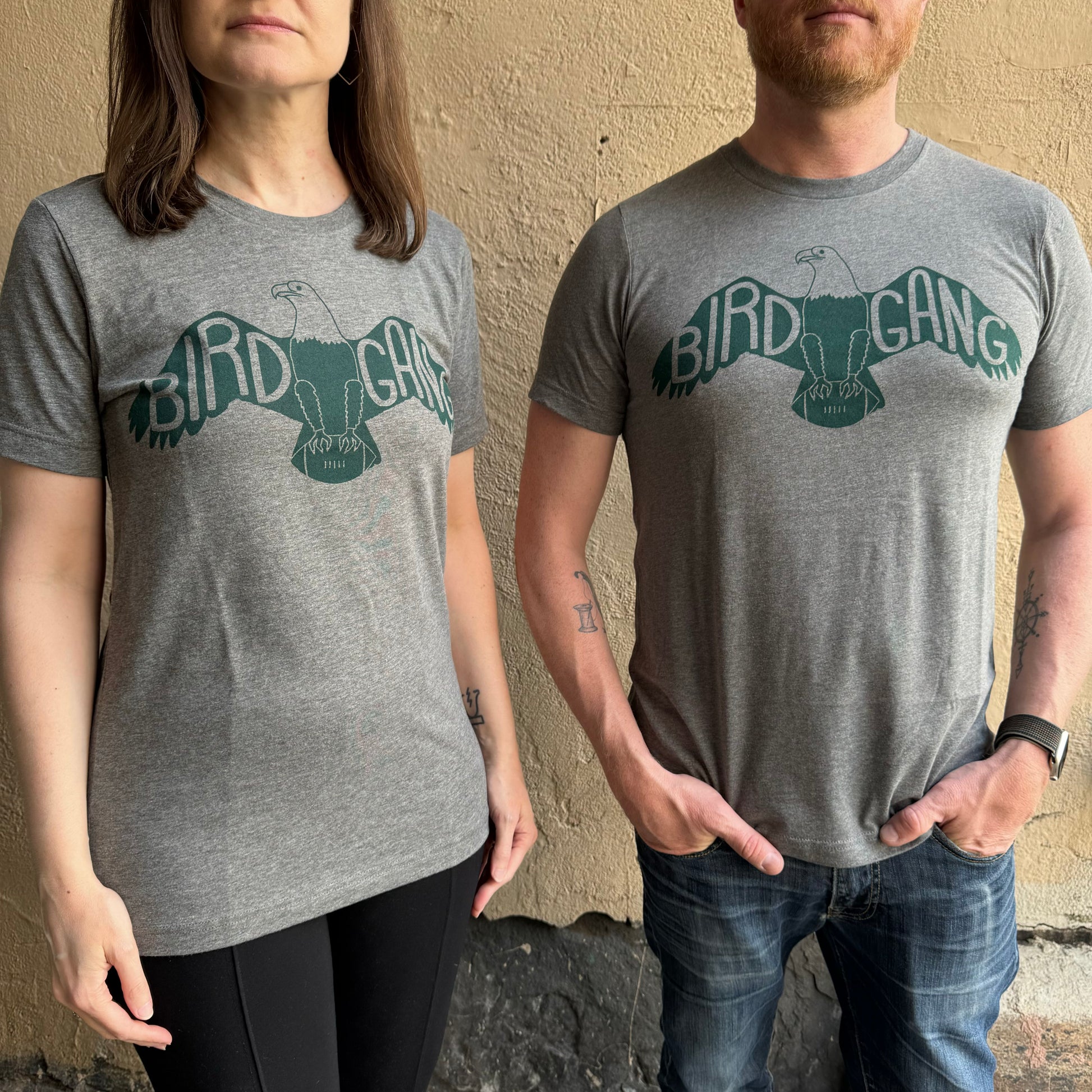 Two people stand against a beige wall wearing matching grey Bird Gang T-Shirts with an eagle graphic and the text "Philly’s Bird Gang." The unisex exit343design t-shirts fit them well. The person on the left has longer hair; the one on the right has a beard and crossed arms.