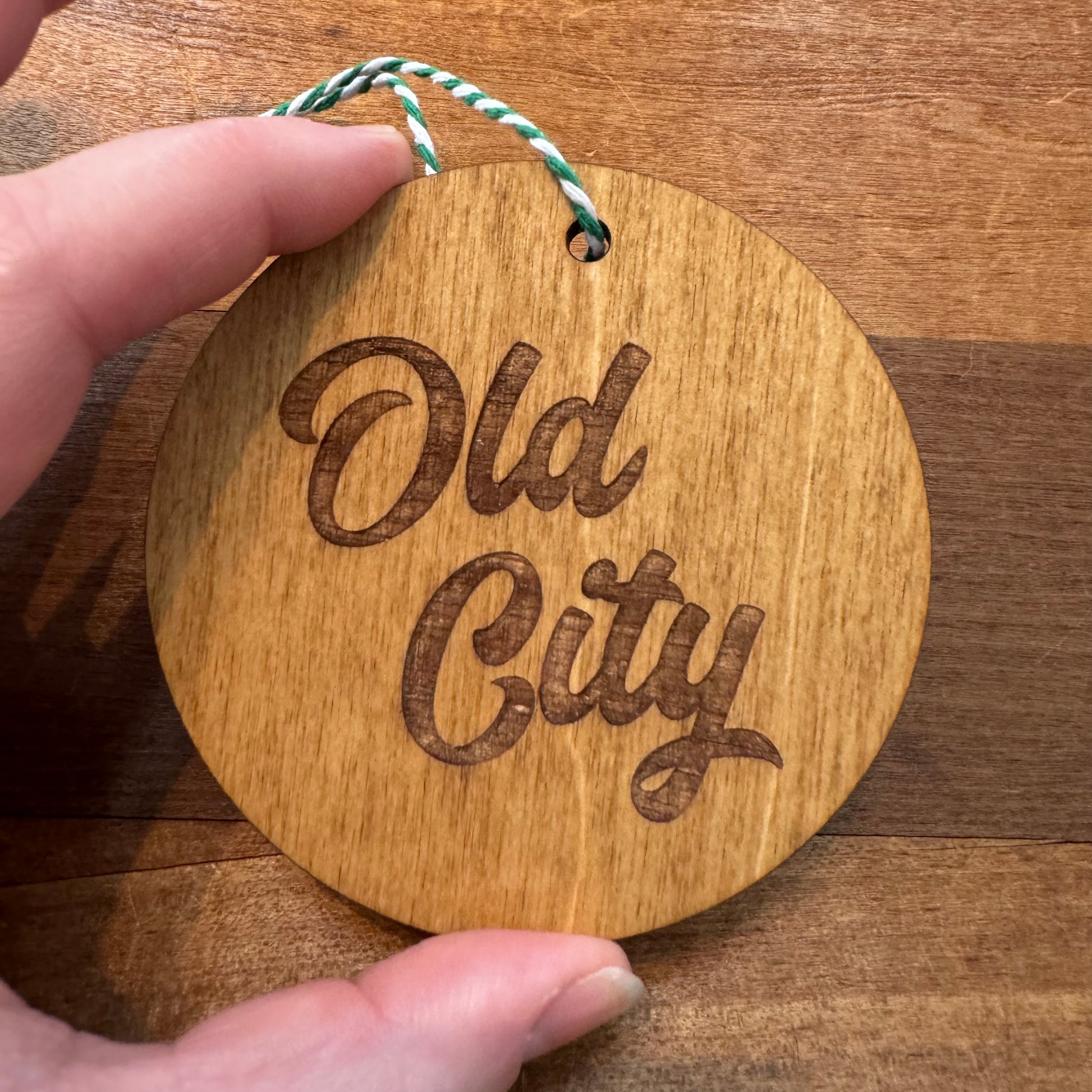 A hand holds a circular wooden ornament from Frankadelphia's Philly Wood Ornaments collection, featuring a laser-etched "Old City" design and attached to a green and white braided string, perfect for anyone who loves Philly-themed decorations.