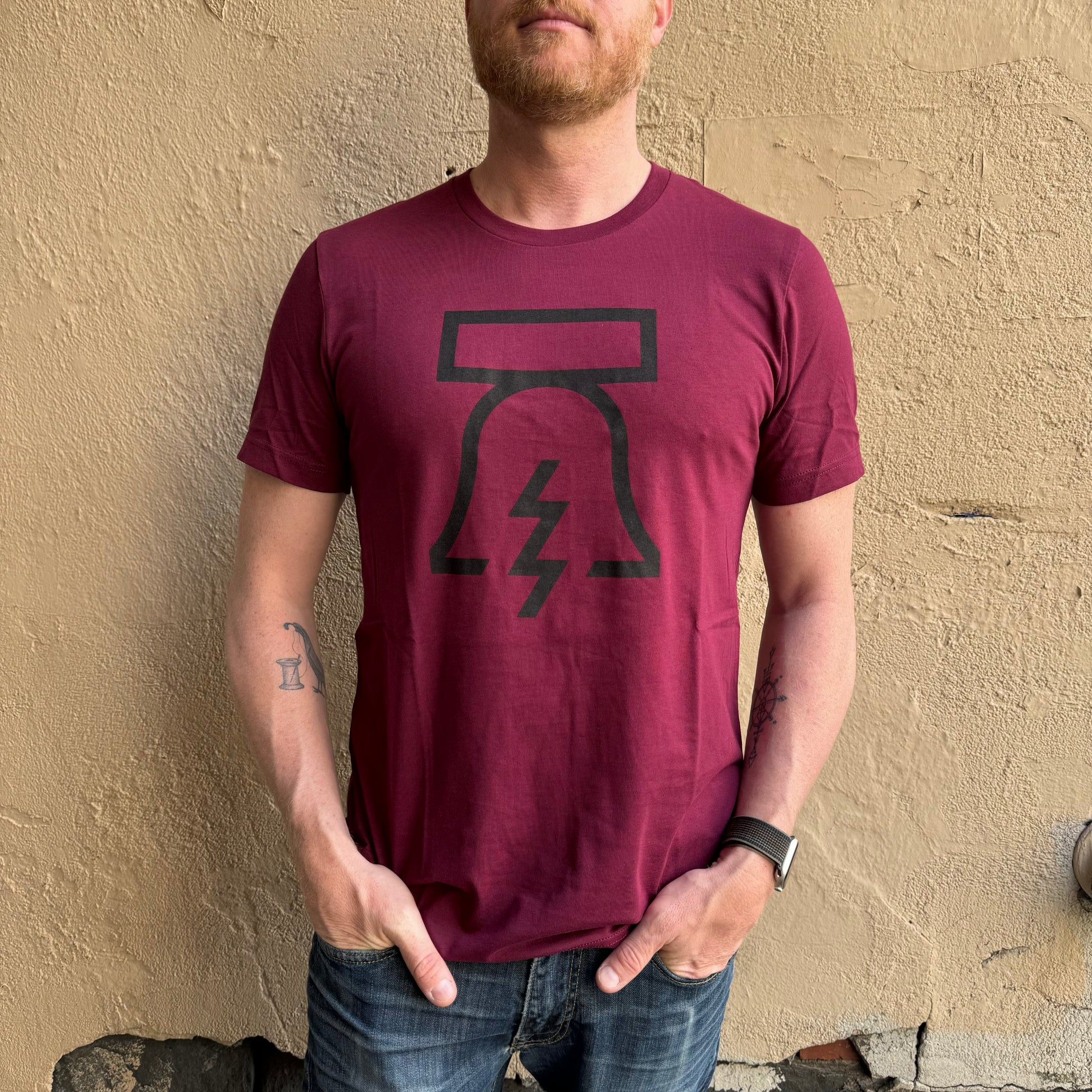 A person with a beard wearing a maroon unisex Philadelphia Independents Bell & Bolt T-Shirt, standing against a textured beige wall.