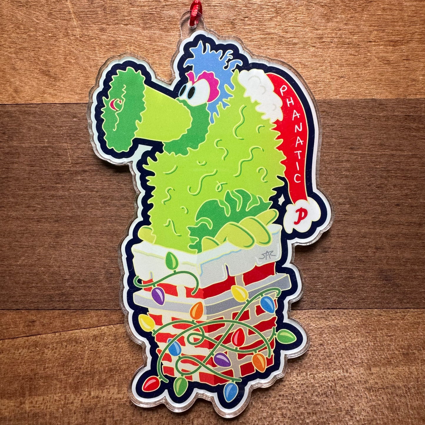 Introducing the Philly Acrylic Ornaments III by Jeffrey Arden Art: a vibrant acrylic ornament featuring a green, fuzzy character called "Phanatic," adorned with a Santa hat and standing on a stack of books wrapped in Christmas lights. This piece is perfect for fans of Philly sports-themed decorations.