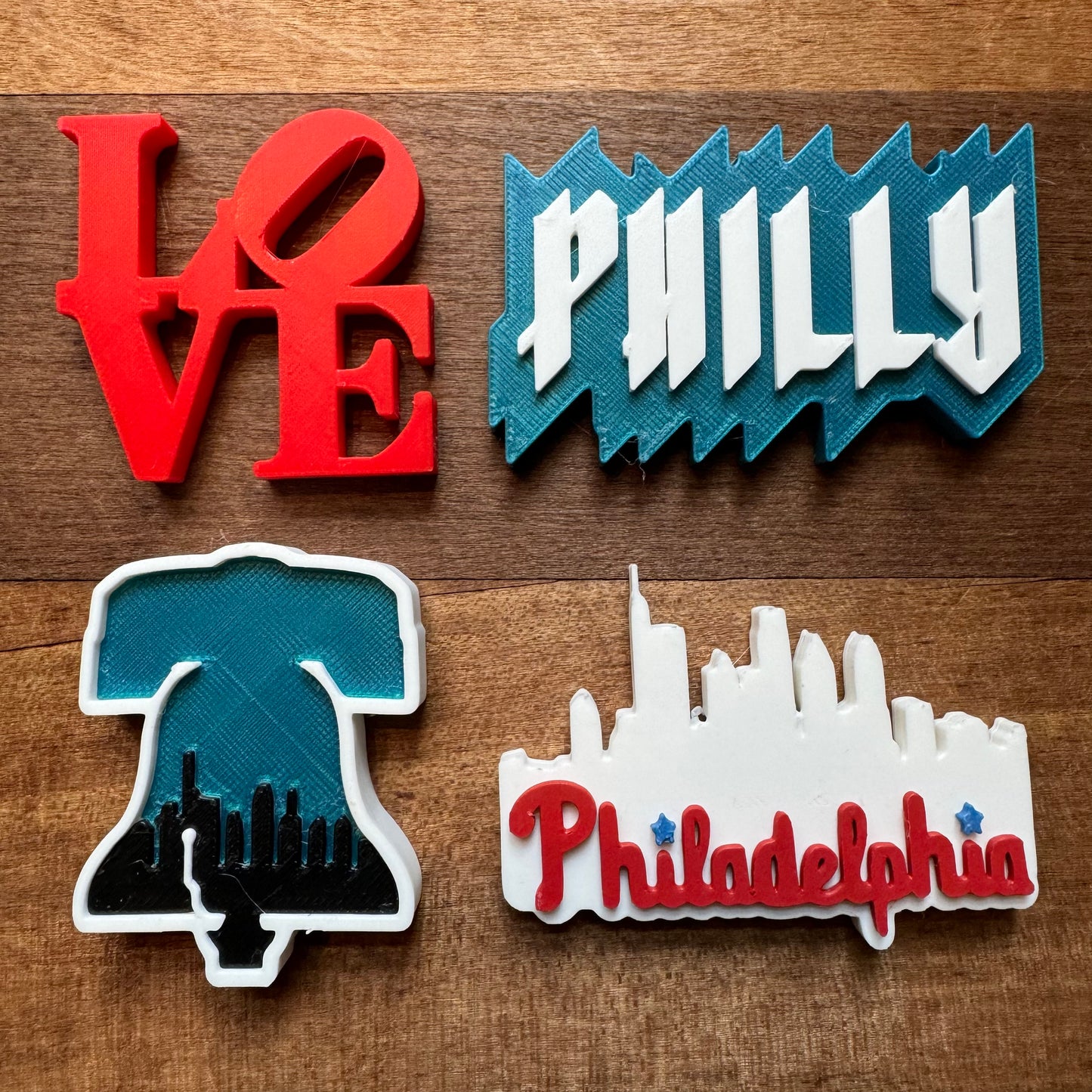 Four Philly-themed magnets on a wooden surface: a red "LOVE" sign, a blue "PHILLY" sign, a black and blue Liberty Bell with skyline, and a white Philadelphia skyline with "Philadelphia" in red text. All pieces are finely crafted 3D-printed designs perfect for any fridge. These Philly Magnets by Rosewood Home add the perfect touch of local pride to your kitchen decor.