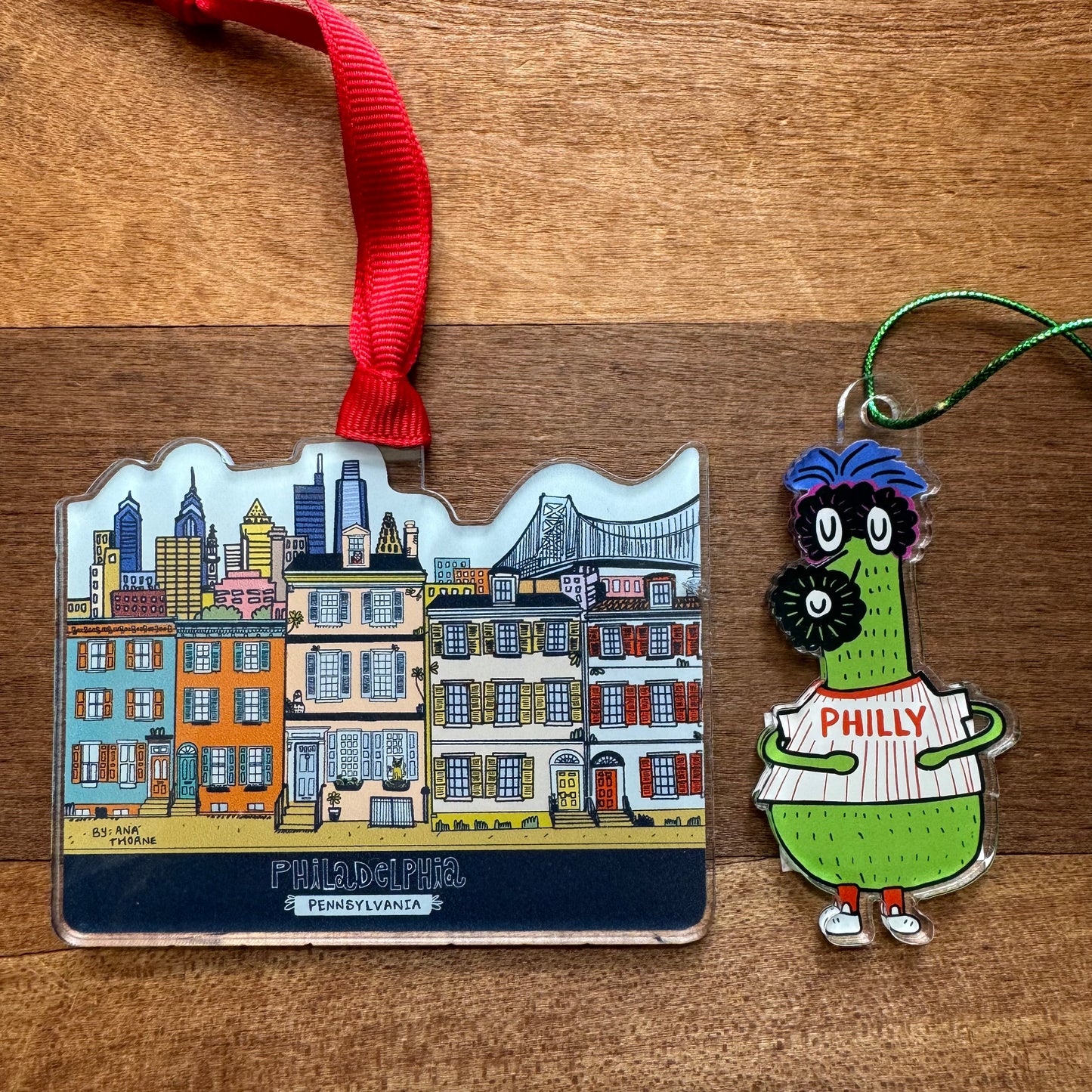 Two Philly Acrylic Ornaments from Ana Thorne: one features a colorful illustration of Philadelphia's skyline and landmarks, while the other showcases a green character wearing a "Philly" baseball shirt, embodying true Philly pride. Both lie on a wooden surface, capturing the essence of Philly Rowhome charm.