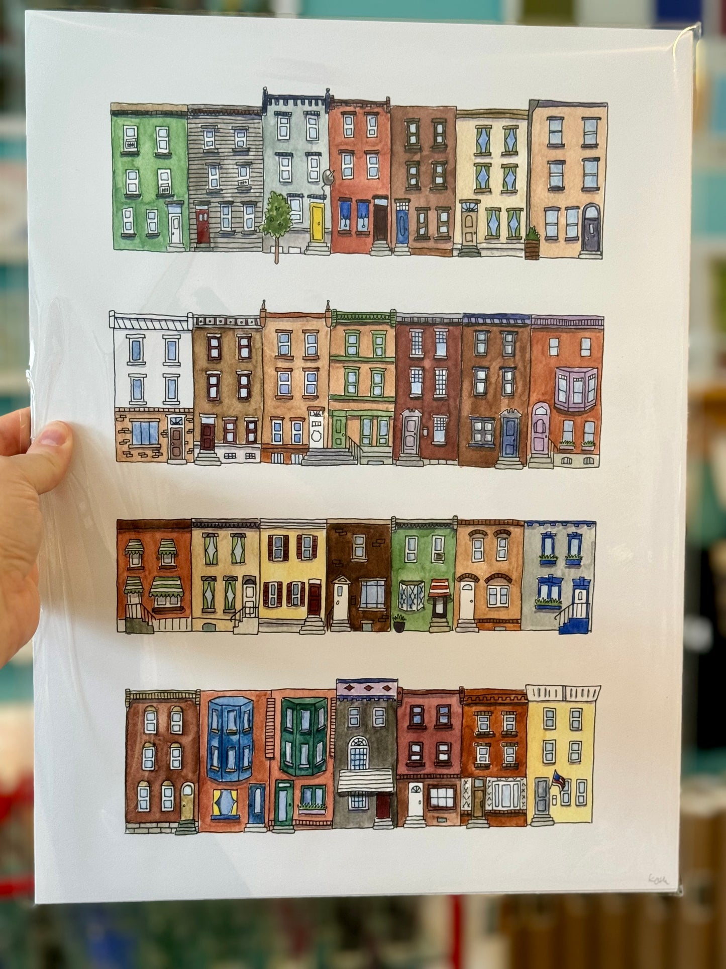 A person holds up a "Philadelphia Prints" illustrated print by Kate Otte, featuring four rows of colorful, stylized townhouses and a hint of Philly Lingo.