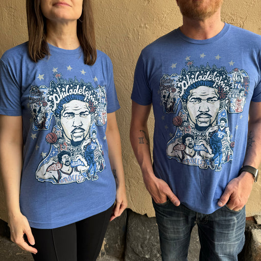 Two individuals wearing blue unisex Paul Carpenter T-shirts with Philadelphia-themed graphics featuring a basketball player and various related images. The shirts have a detailed and colorful design, a nod to Joel Embiid's electrifying presence on the Sixers court.