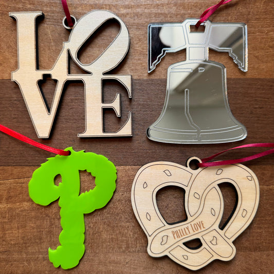 A set of four intricately designed Laser Cut Ornaments from Rosewood Home is displayed on a wooden surface, featuring the word "LOVE," a bell, a pretzel inscribed with "Philly Love," and a green emblem resembling the letter "P.