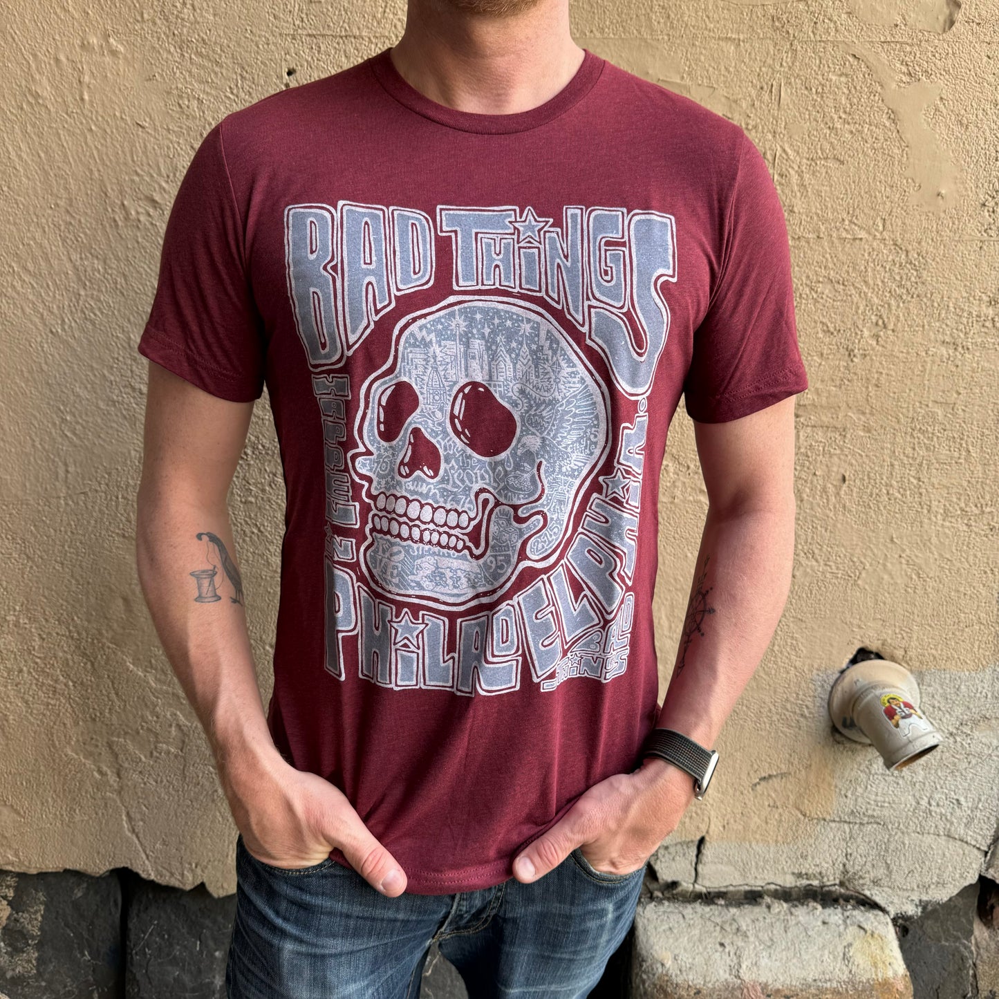 Person wearing a super soft unisex maroon Bad Things Happen T-Shirt by Paul Carpenter with a graphic of a skull and the text "Bad Things Happen in Philadelphia," standing against a beige wall.