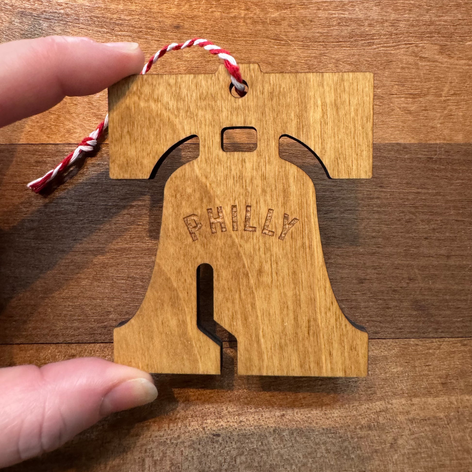 A hand holds a Frankadelphia Philly Wood Ornament, shaped like the Liberty Bell with "Philly" engraved on it. This charming piece of Philly-themed decor features a red and white string for hanging, perfect for adding a touch of Old City history to your collection.