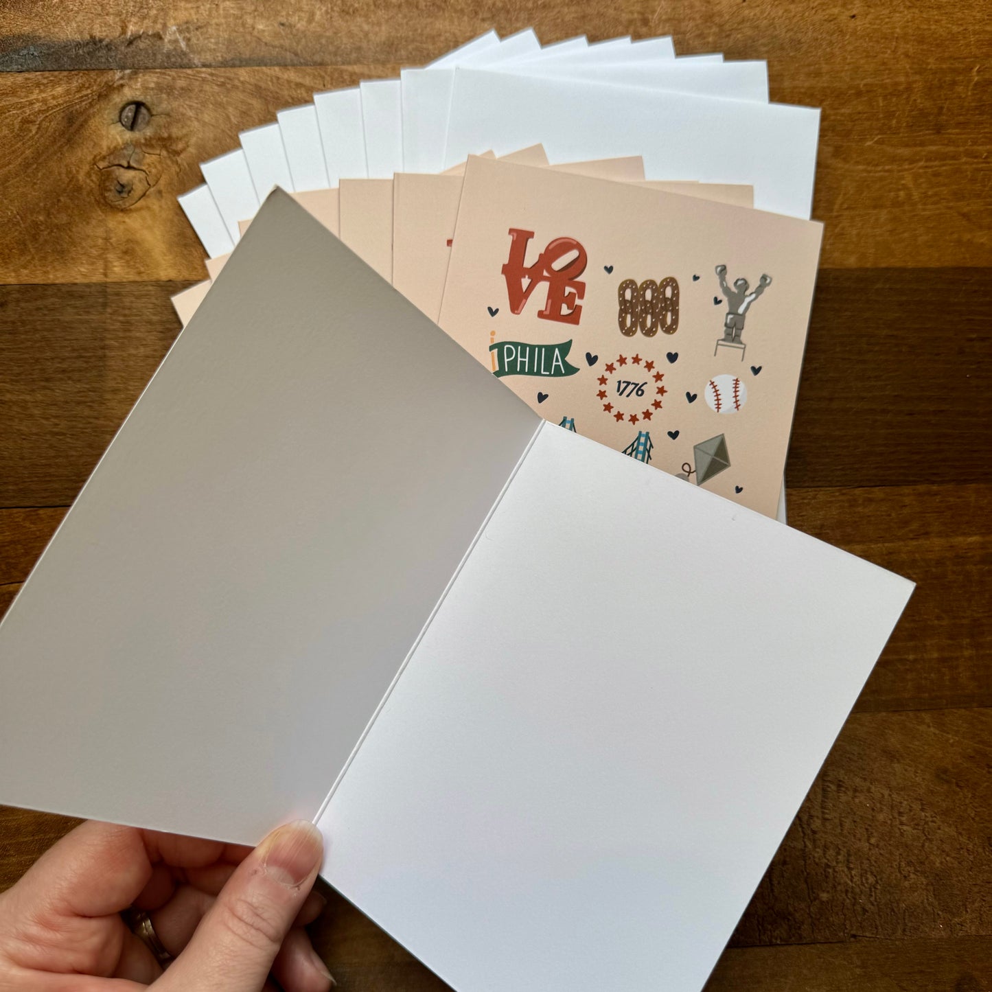 A hand is holding open an unmarked greeting card next to a stack of Parcel Island's Philadelphia Notecard Pack, showcasing various Philadelphia-themed designs, all laid out on a wooden surface.