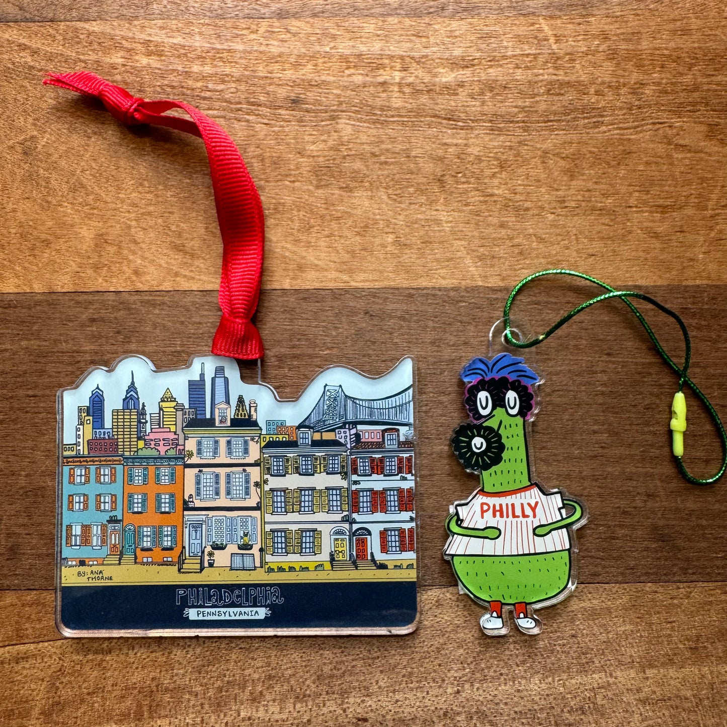 Two Philly Acrylic Ornaments by Ana Thorne rest on a wooden surface: one showcases a vibrant cityscape with famous landmarks, embodying ultimate Philadelphia pride, while the other features a charismatic mascot holding a sign that reads "Philly.