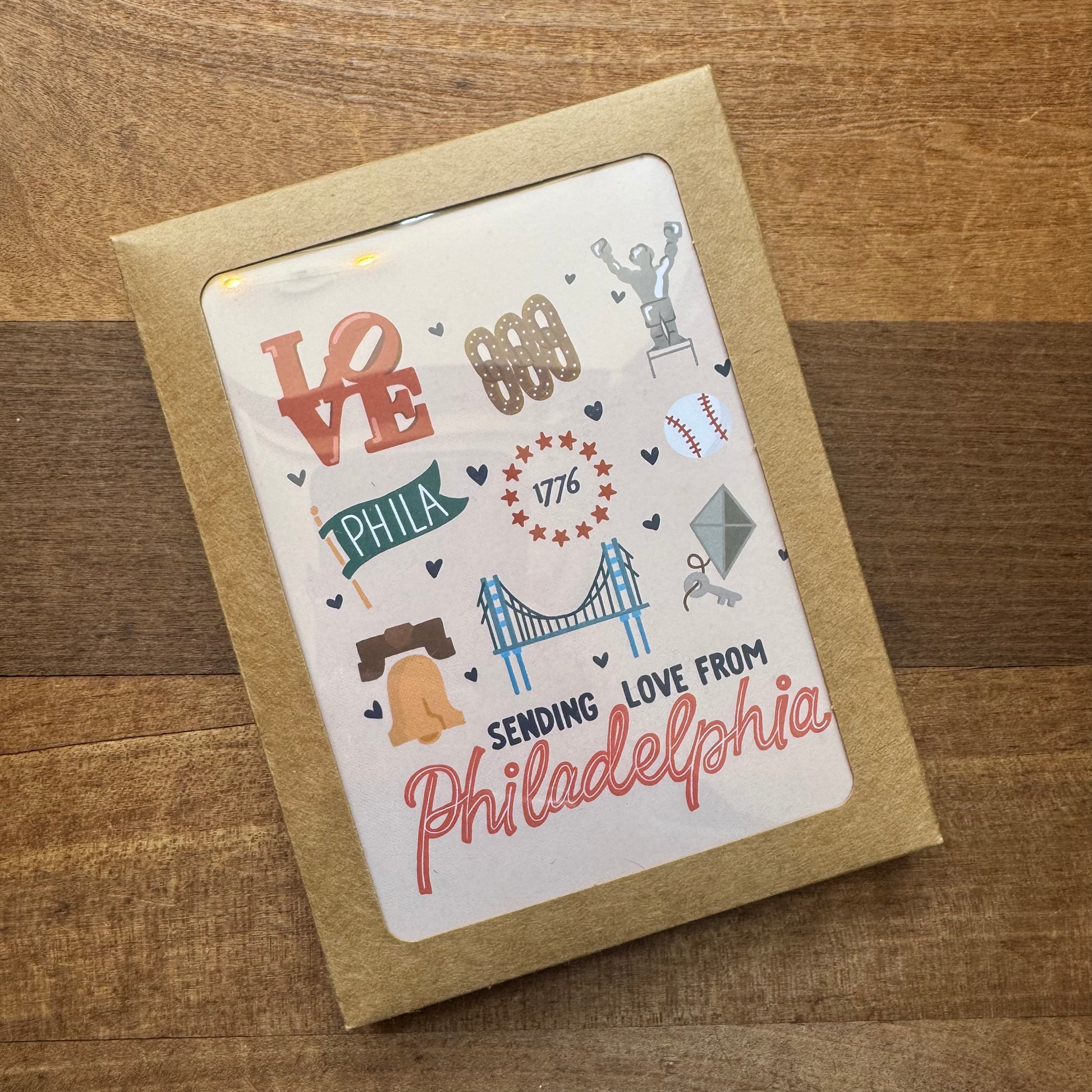 A postcard from the Parcel Island Philadelphia Notecard Pack, showcasing iconic Philadelphia symbols such as the Liberty Bell, the LOVE sculpture, 1776 stars, a bridge, and the text "Sending Love from Philadelphia," is placed on a wooden surface. This charming piece truly captures the essence of Philly phlair.