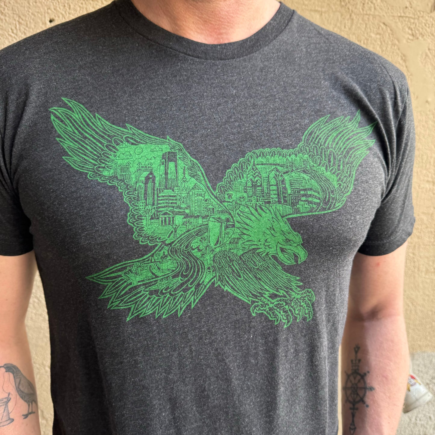 A person wearing a dark gray T-shirt with a green illustration of an eagle flying over a cityscape on the chest, reminiscent of Paul Carpenter's Swooping Eagle T-Shirt. They also have tattoos on their left arm.