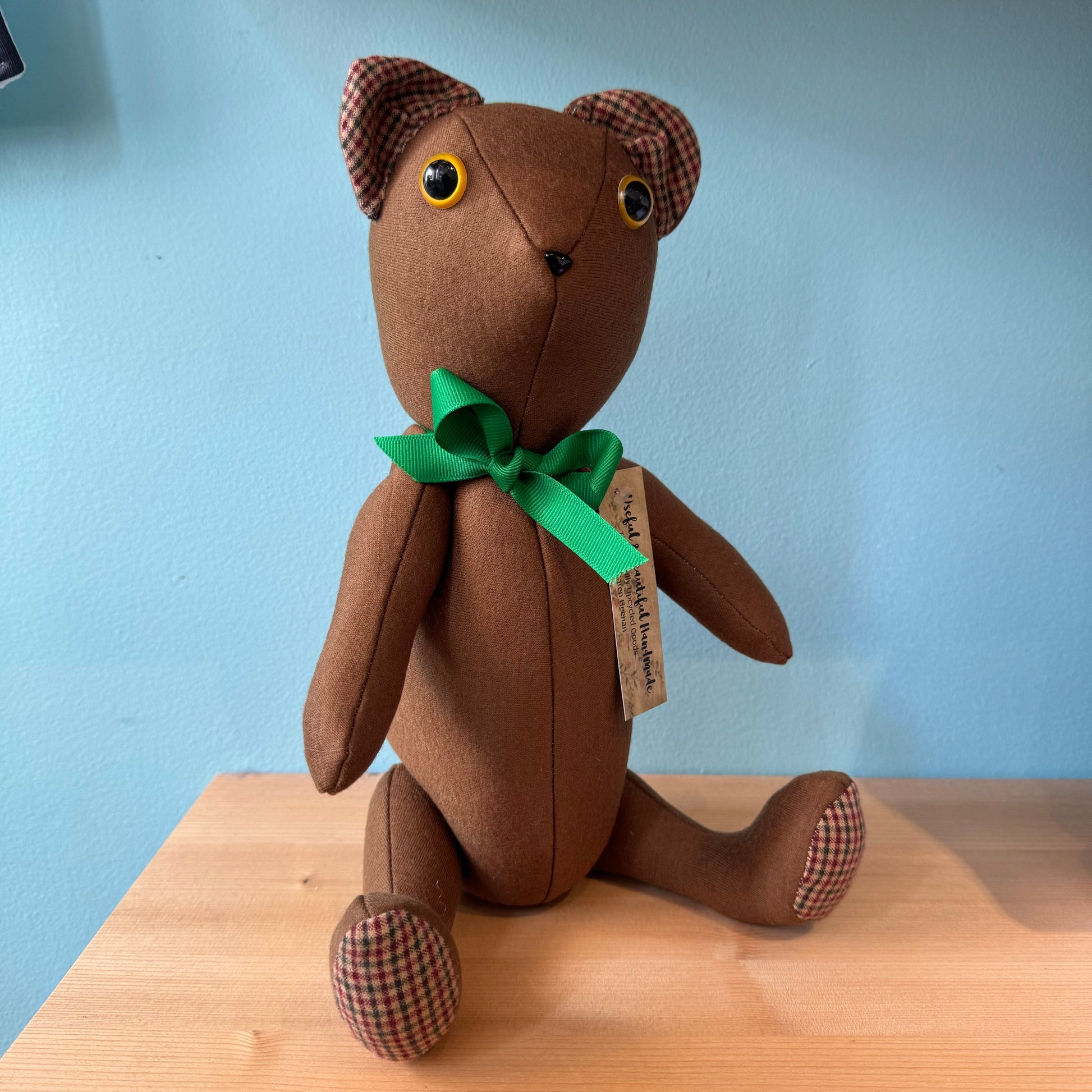 A charming brown teddy bear from Useful & Beautiful Handmade's Stuffed Teddy Bears collection sits on a wooden surface against a light blue background. The bear, with plaid ears and feet, is crafted from upcycled materials and wears a green bow with a tag attached, making it an ideal baby gift.