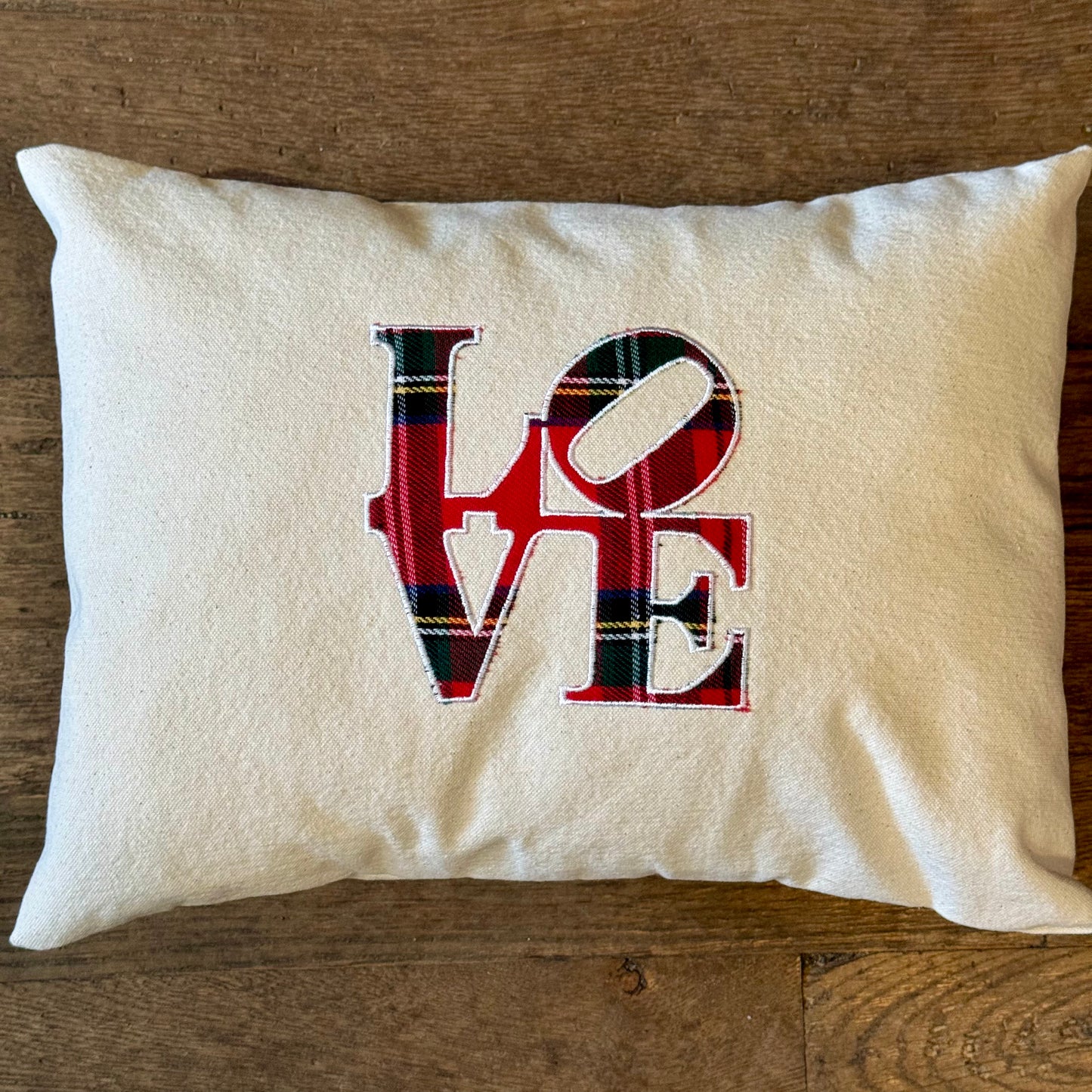 A rectangular white pillow from The Pillow Works' Holiday Plaid Pillows collection, featuring the word "LOVE" printed in red and green plaid letters on the front, exudes festive charm as it rests gracefully on a wooden surface.