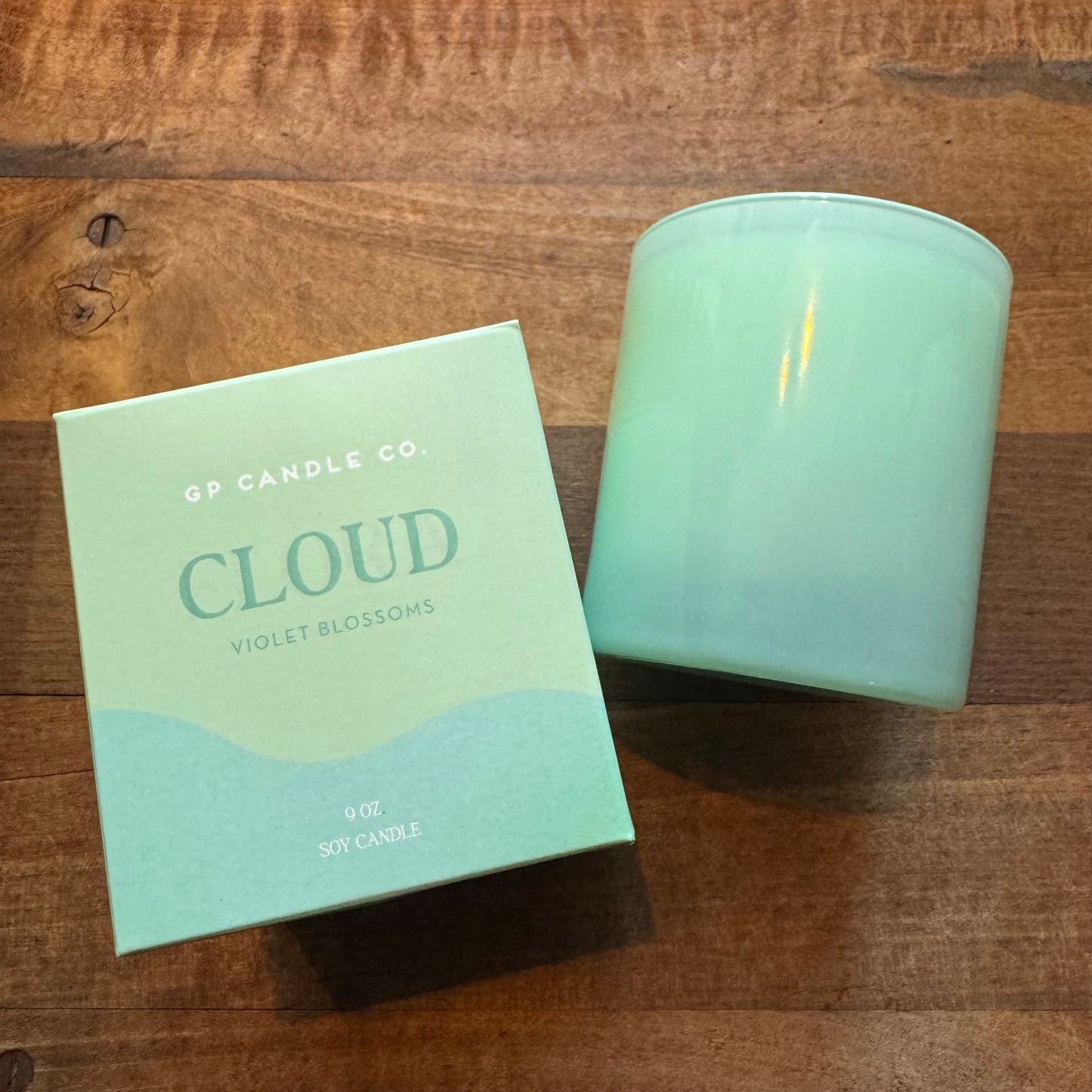 A hand-poured Soy Candles-Hue Collection candle named "cloud violet blossoms" by GP Candle Co beside its packaging box on a wooden surface.