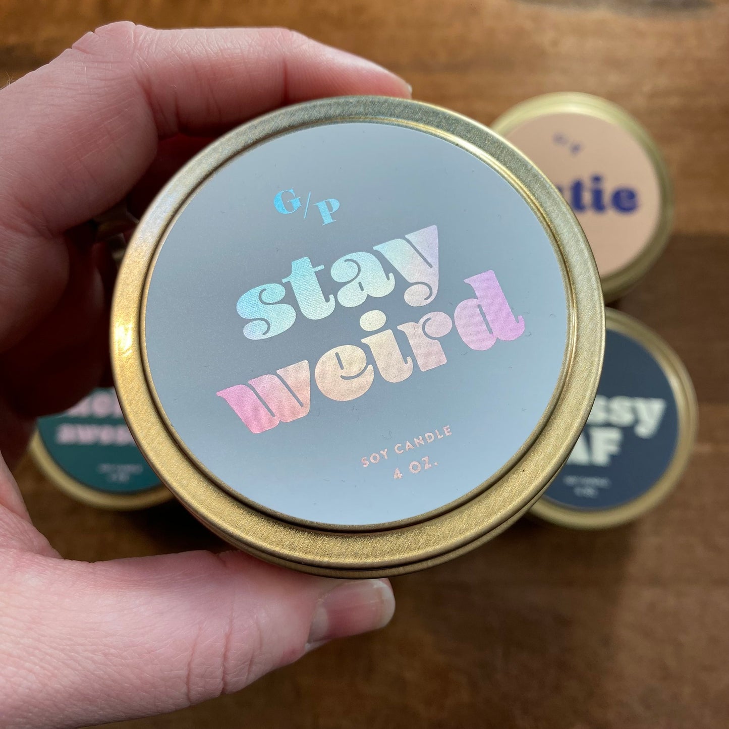 A hand holding a small, GP Candle Co Tin Collection Soy Candles II with the phrase "stay weird" on the lid.