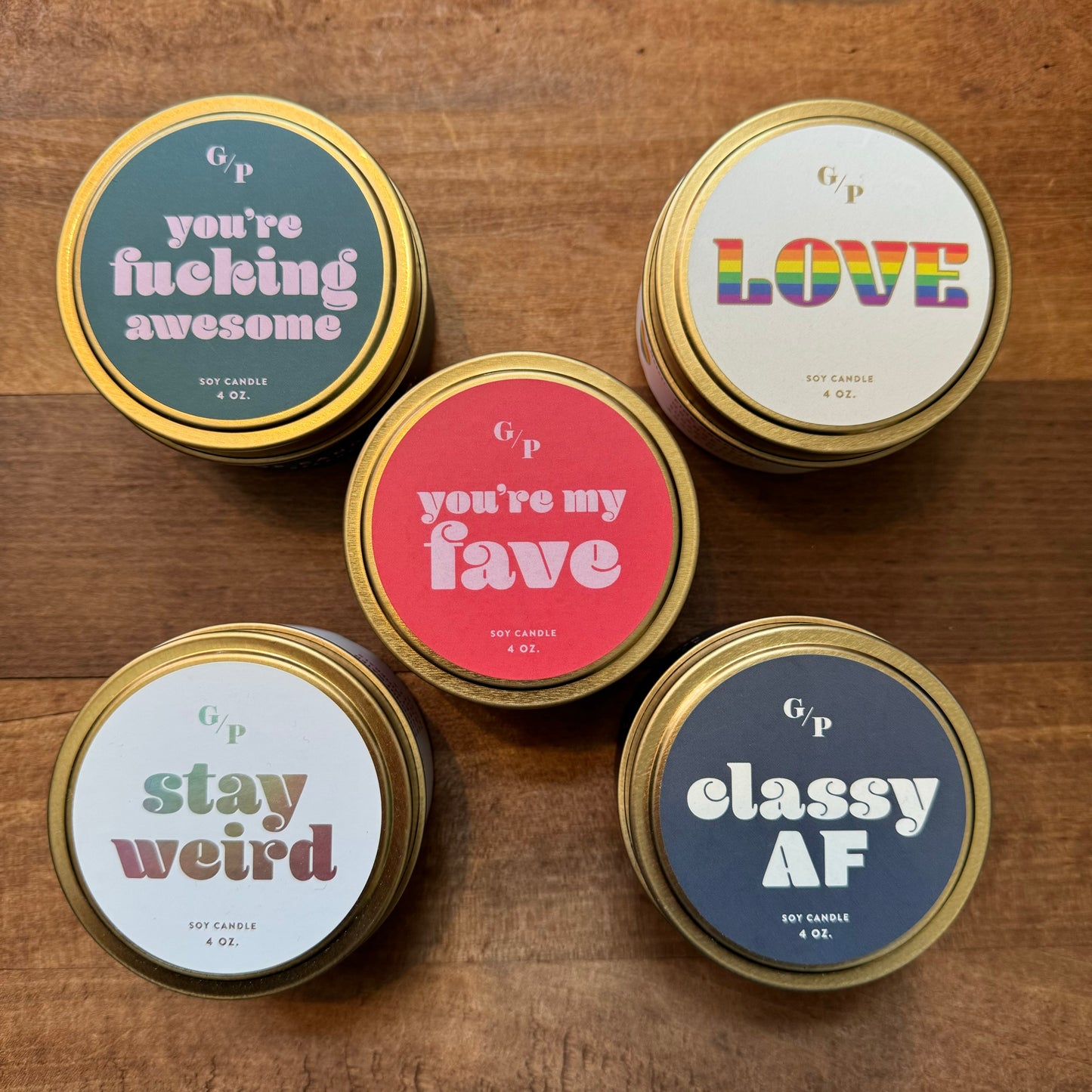 Six Tin Collection Soy Candles II from GP Candle Co with playful phrases on the labels arranged on a wooden surface.