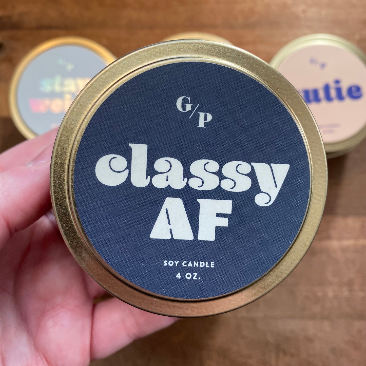 A hand holding a 4 oz Tin Collection Soy Candles II from GP Candle Co with "classy af" labeled on the lid.