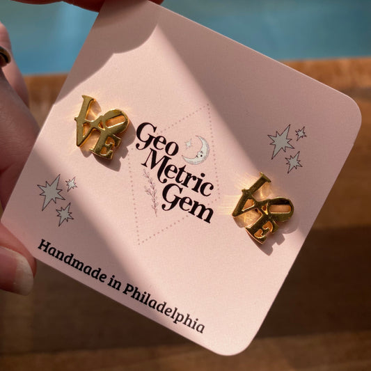 A pair of gold-plated, bow-shaped LOVE Cutout Earrings for sensitive ears displayed on a pink card with GeoMetricGem branding.