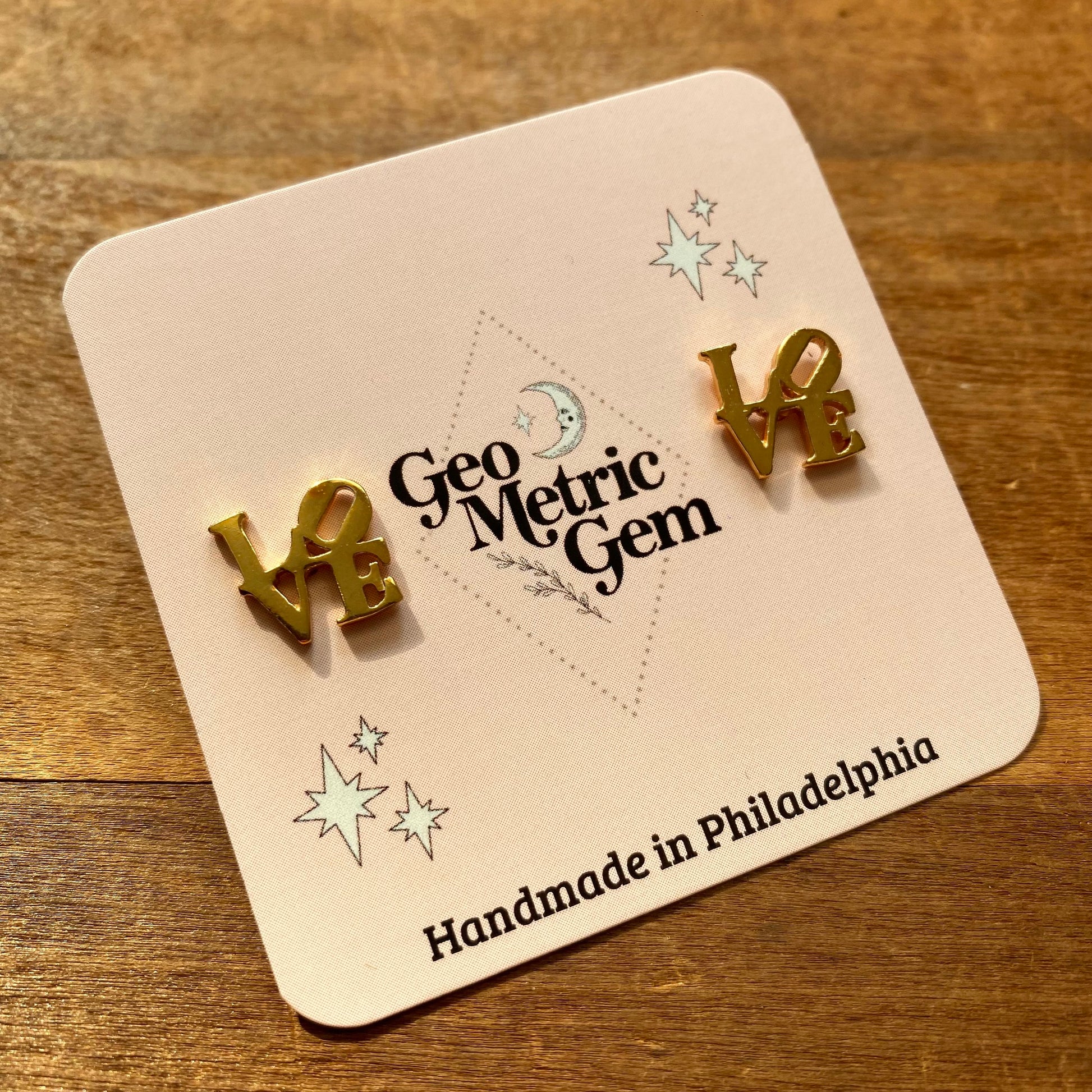 Two gold plated "LOVE" shaped earrings on a display card for GeoMetricGem, handmade in Philadelphia.