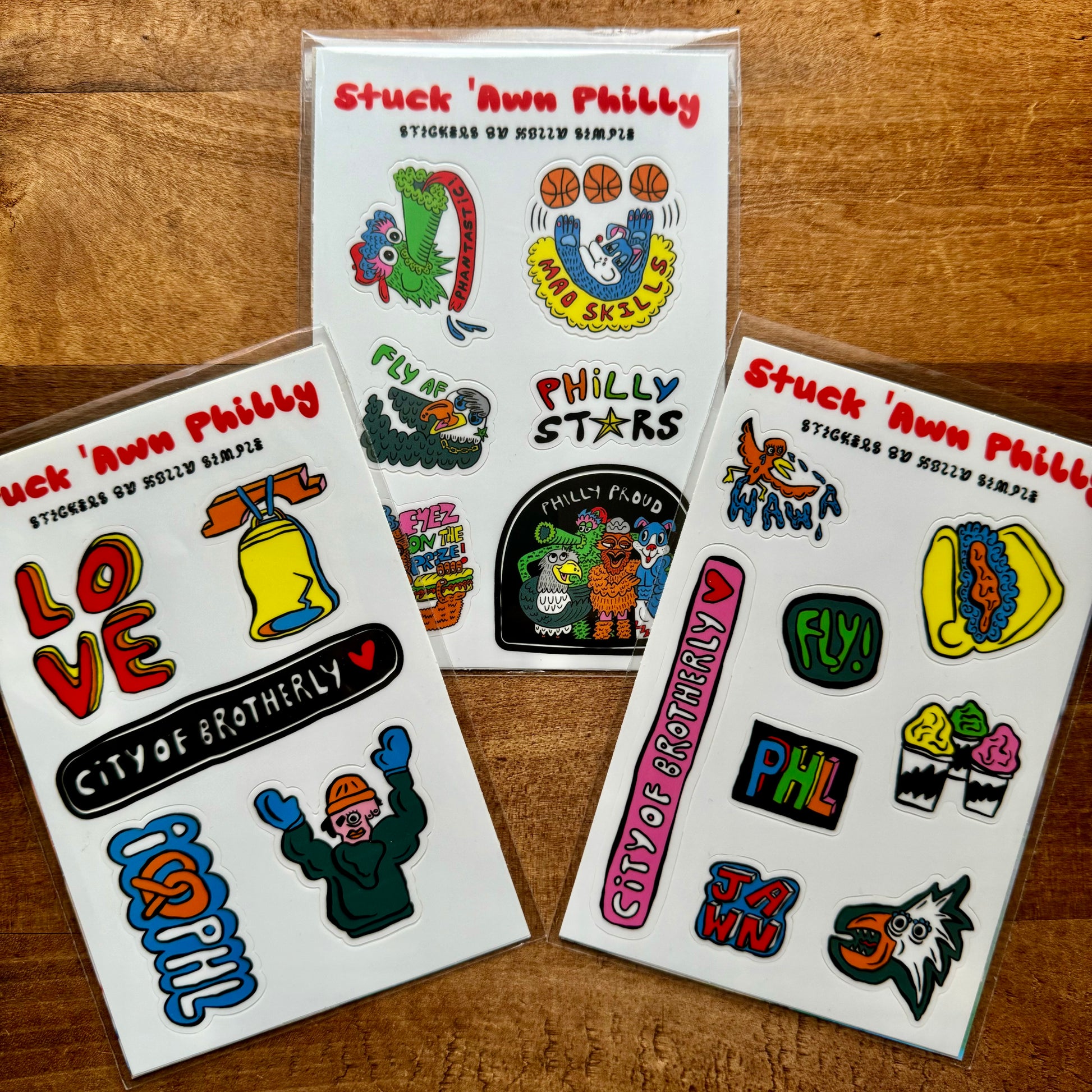 Three packs of Philly Sticker Packs by Holly Simple displayed on a wooden surface. Titled "Stuck Inna Philly," the designs feature local icons, phrases like "City of Brotherly Love," and images such as the Liberty Bell and sports references.