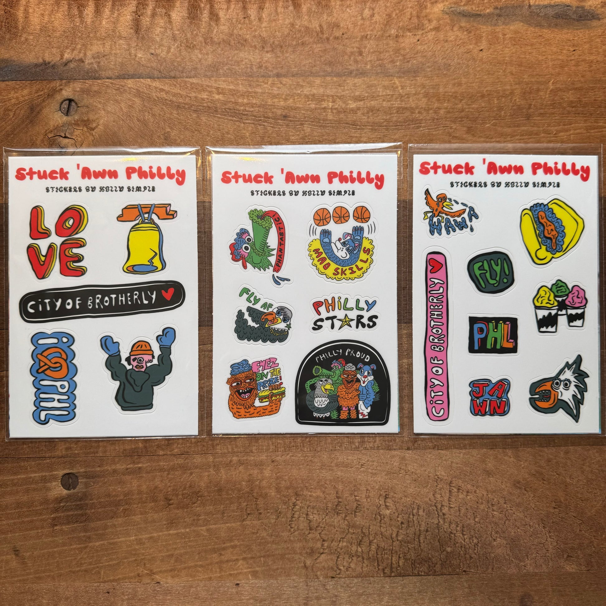 Three packs of "Philly Sticker Packs" by Holly Simple rest on a wooden surface. Each pack includes an array of colorful Philly-themed designs, featuring text, icons, and illustrations related to Philadelphia.