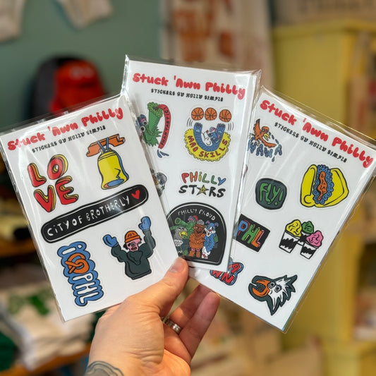 A hand holding three packs of Holly Simple's Philly Sticker Packs, which showcase various designs such as LOVE, sports, food, and city landmarks.