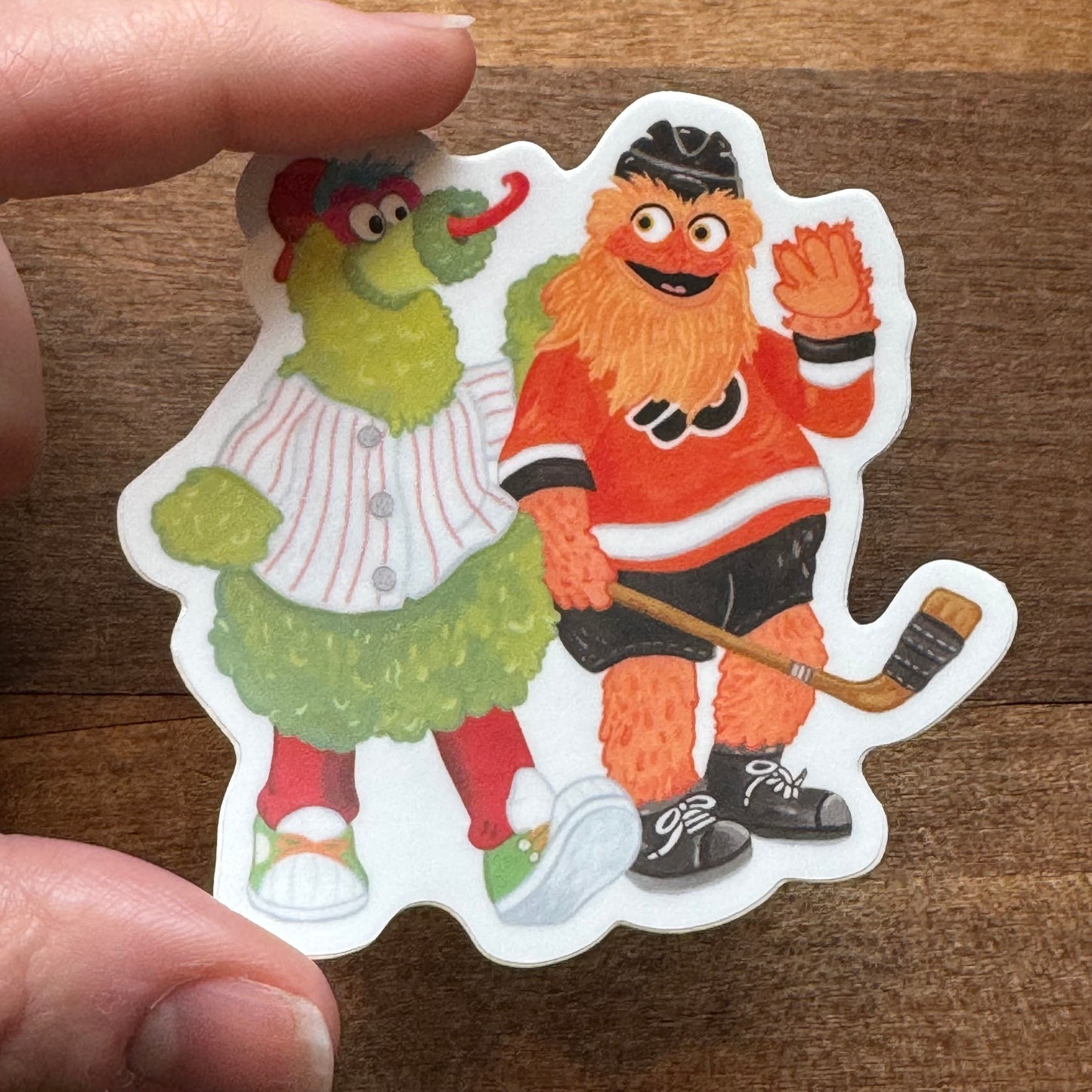 A hand holding a Jamie Bendas Gritty & Phanatic BFF Sticker featuring illustrated characters, one dressed as a baseball player and the other as a hockey player.