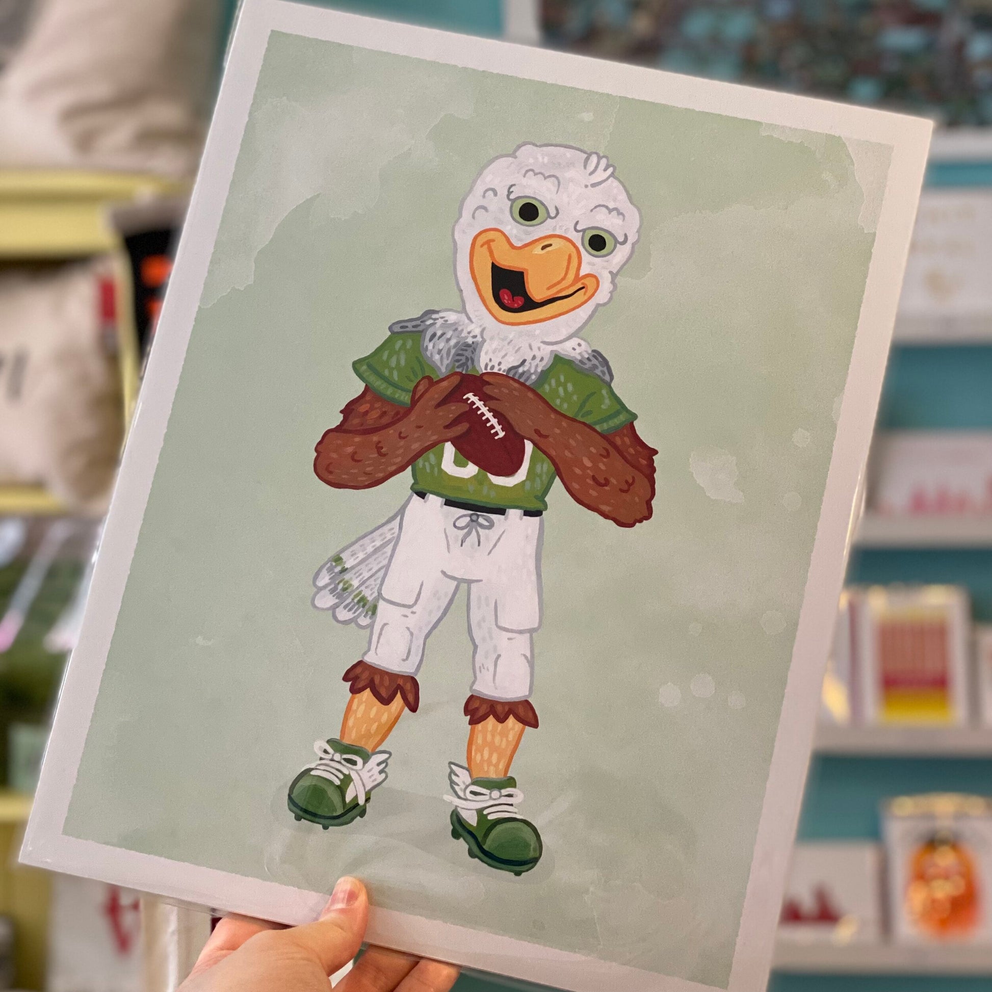 An illustration of Philly Mascot Prints, an anthropomorphic bird dressed in football attire and holding a football, displayed on an archival digital print held by Jamie Bendas.