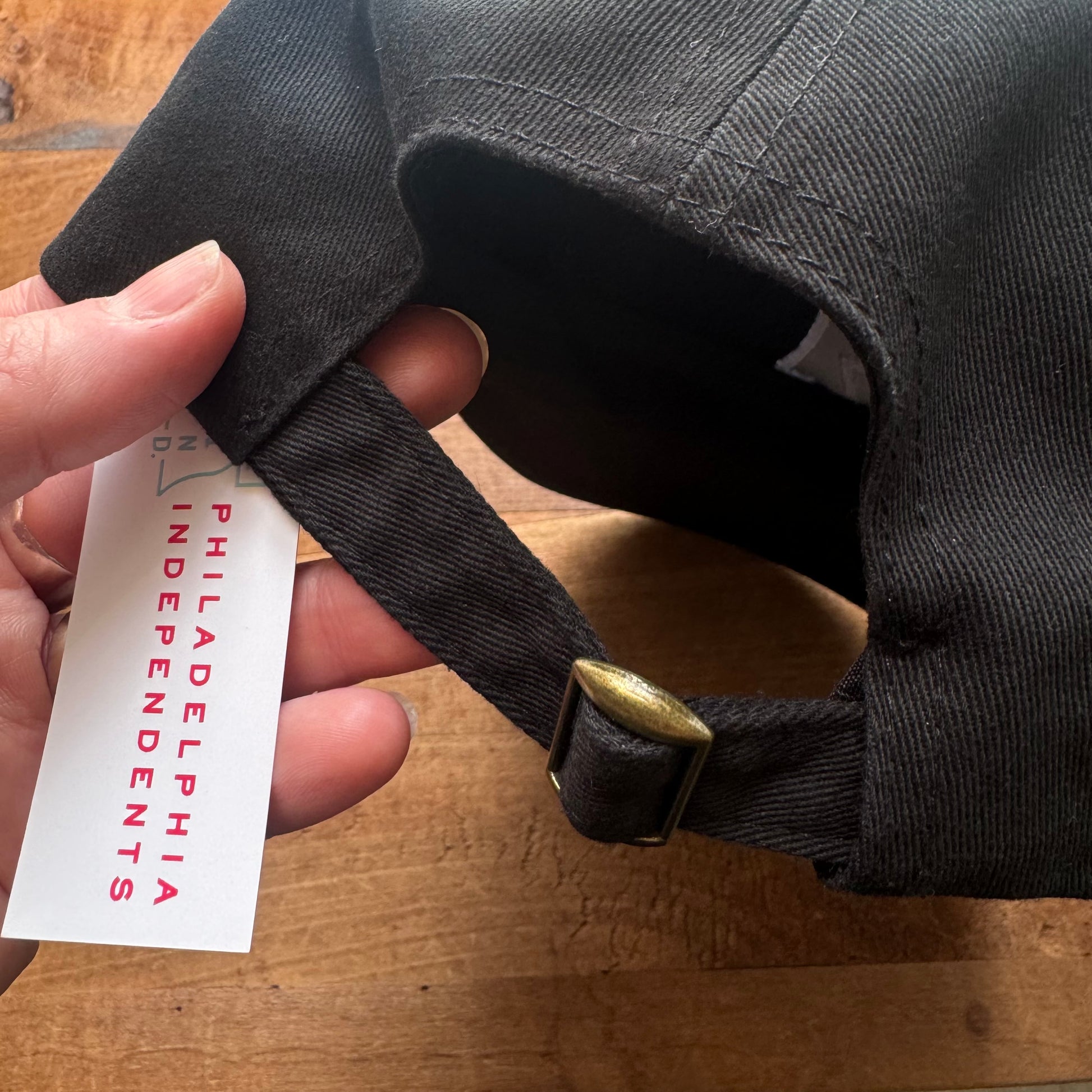 A hand holding a Bell & Bolt Baseball Hat with a "Philadelphia Independents" tag and an adjustable brass buckle strap on a wooden table.