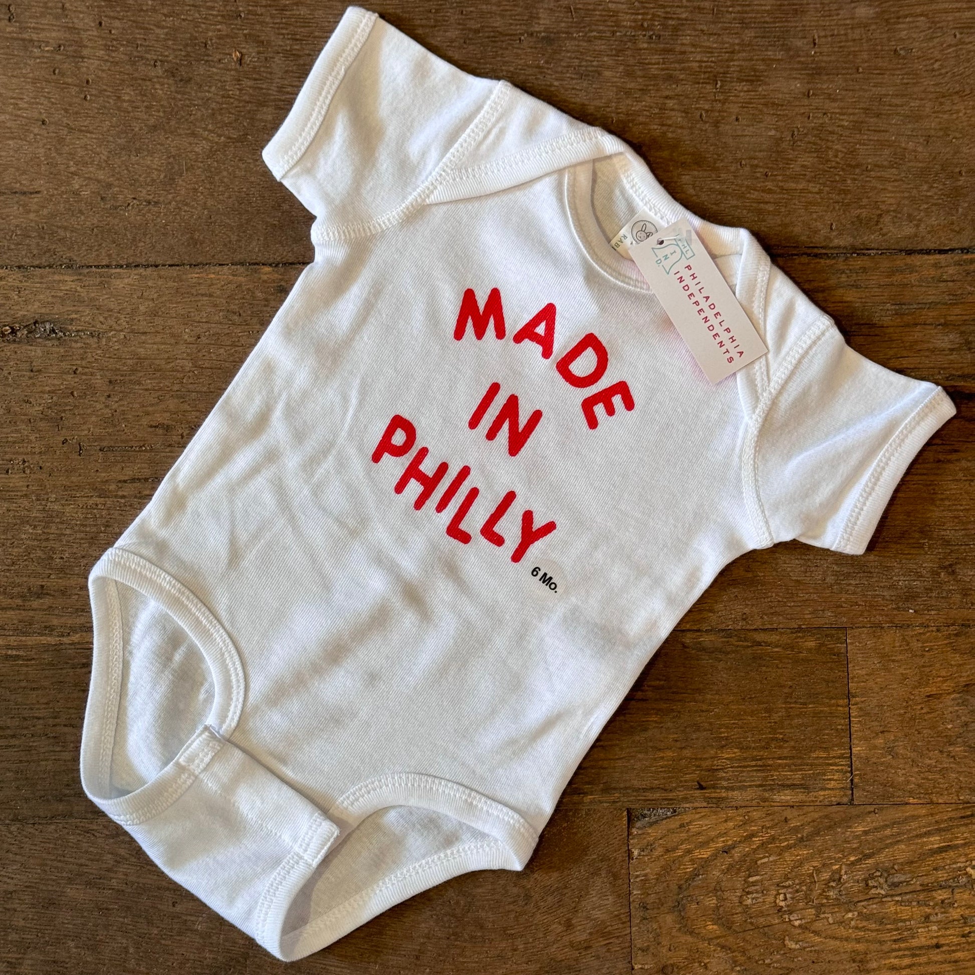 A white baby onesie made of 100% cotton with the Philadelphia Independents Made in Philly Baby Onesie in red letters, displayed on a wooden surface.