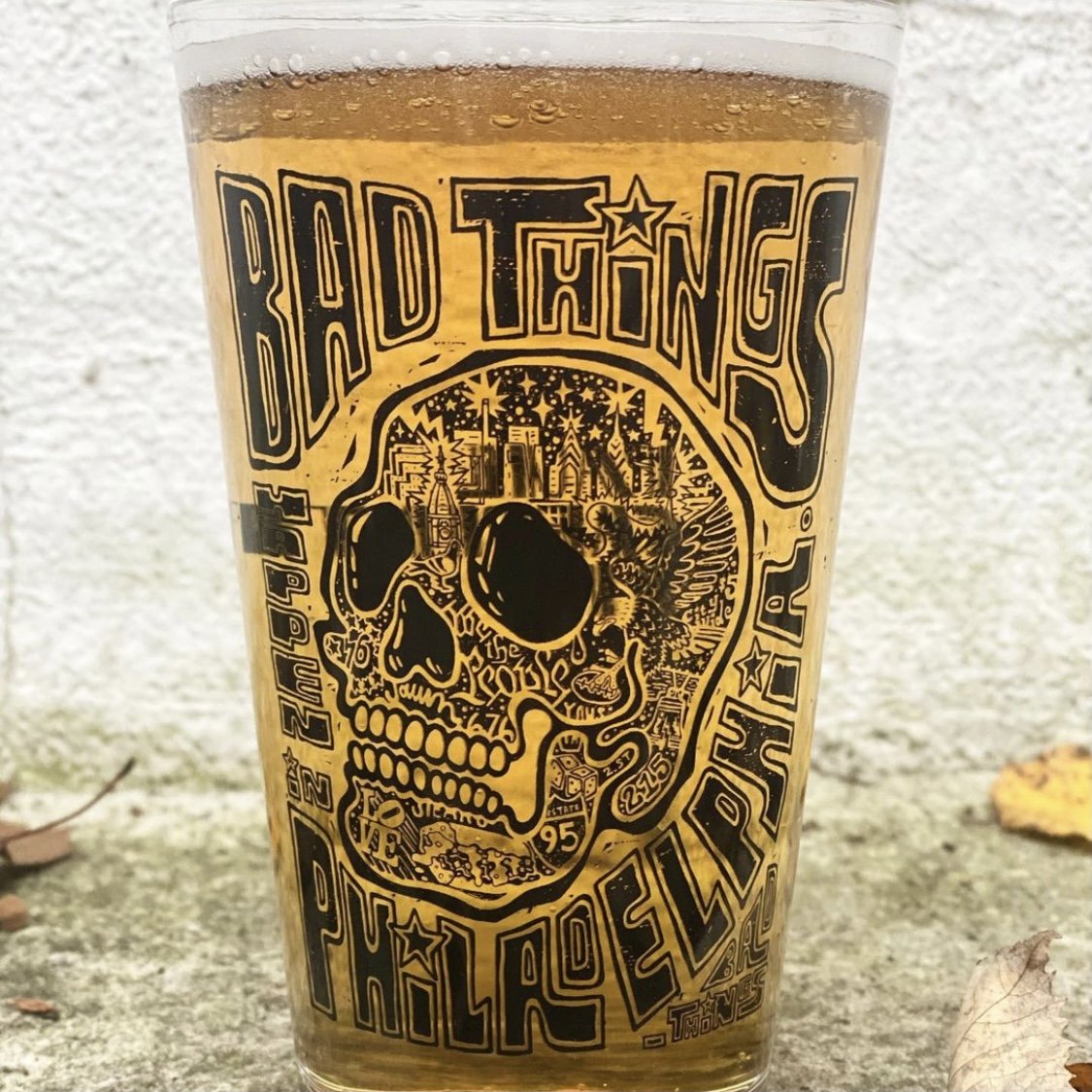 A Philly Pint Glass filled with a golden-colored beverage featuring a graphic with the text "bad things Philadelphia" and skyline imagery.
