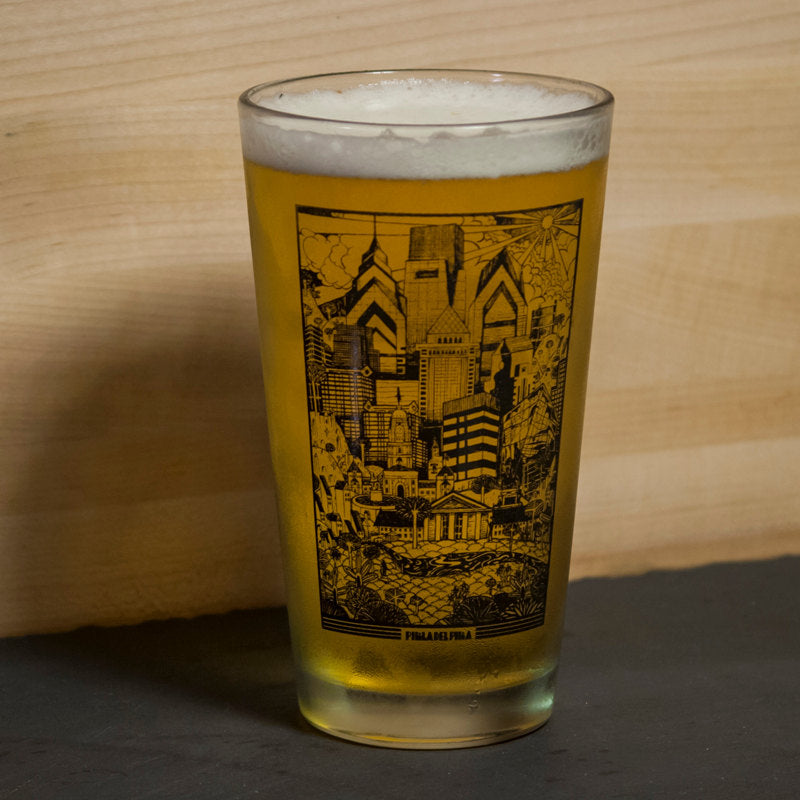 A glass of beer with a Philly Pint Glasses by Paul Carpenter design featuring an illustrated Philadelphia-themed skyline, placed on a wooden surface.