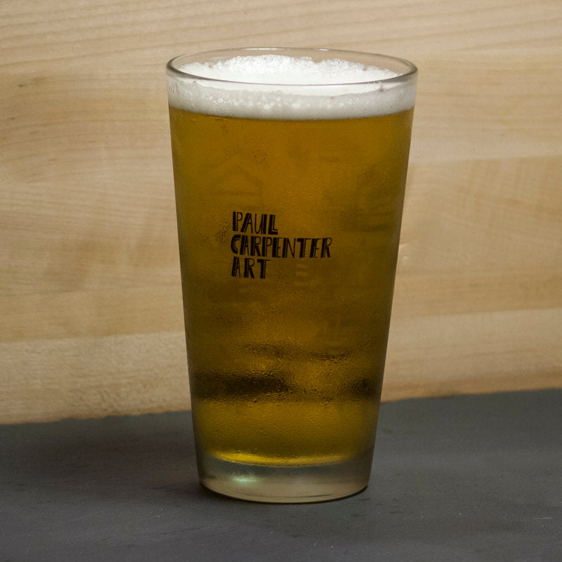 A Philly Pint Glass with foam on top, featuring the text "Paul Carpenter Art" and a Philadelphia-themed skyline on the pint glass, placed on a wooden surface.