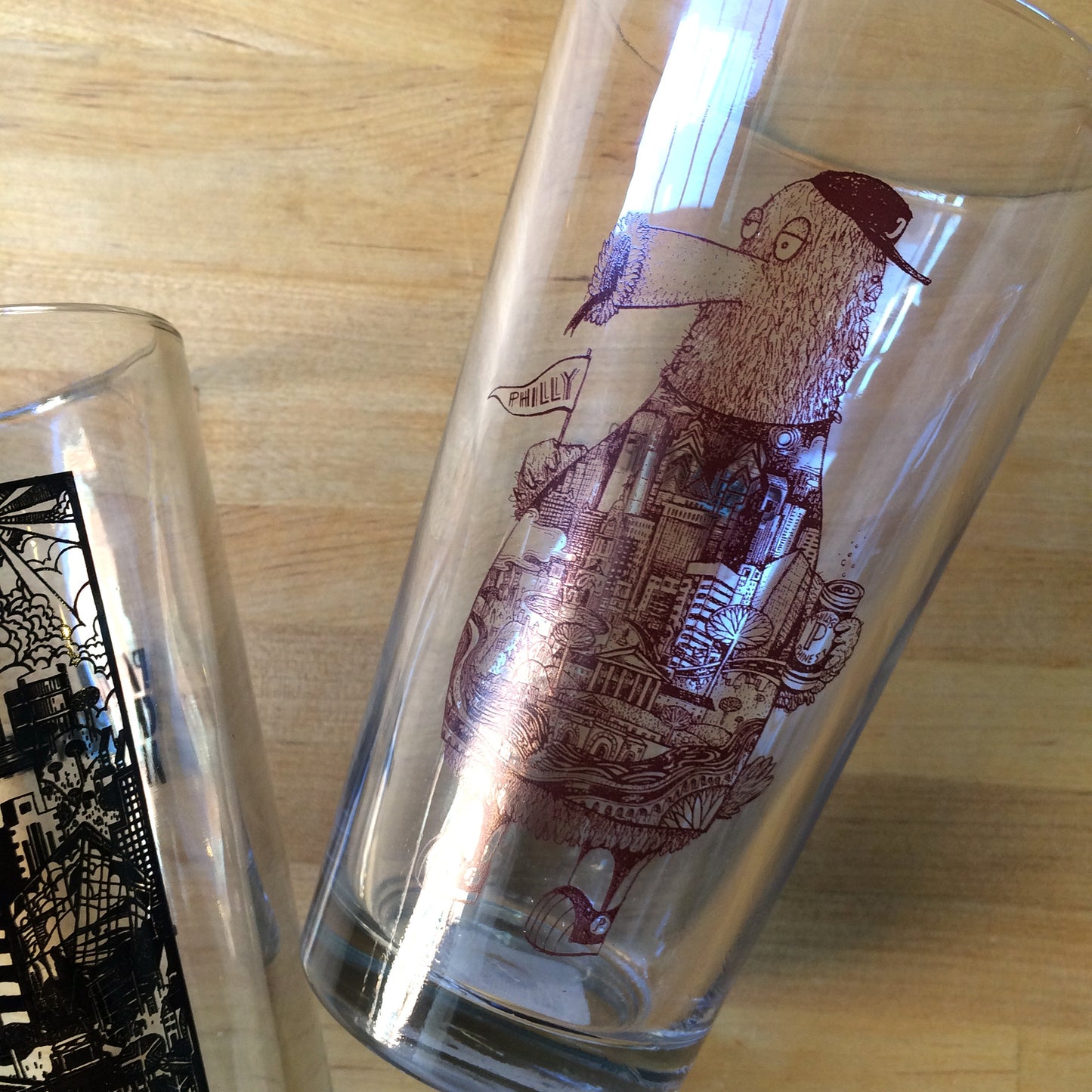 Two decorative Philly Pint Glasses with intricate designs, one featuring a depiction of a character and Philadelphia-themed skyline by Paul Carpenter.