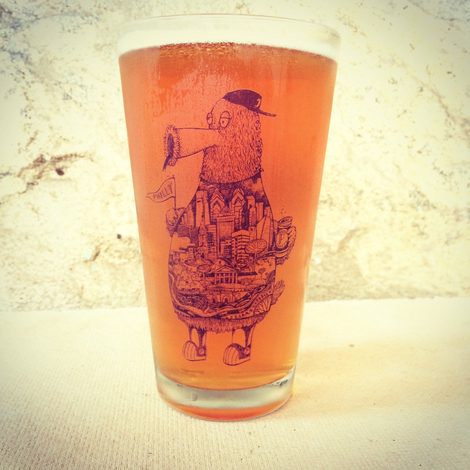 A glass of amber beer in a Philly Pint Glass with an intricate Philadelphia-themed illustration on it, set against a textured backdrop by Paul Carpenter.