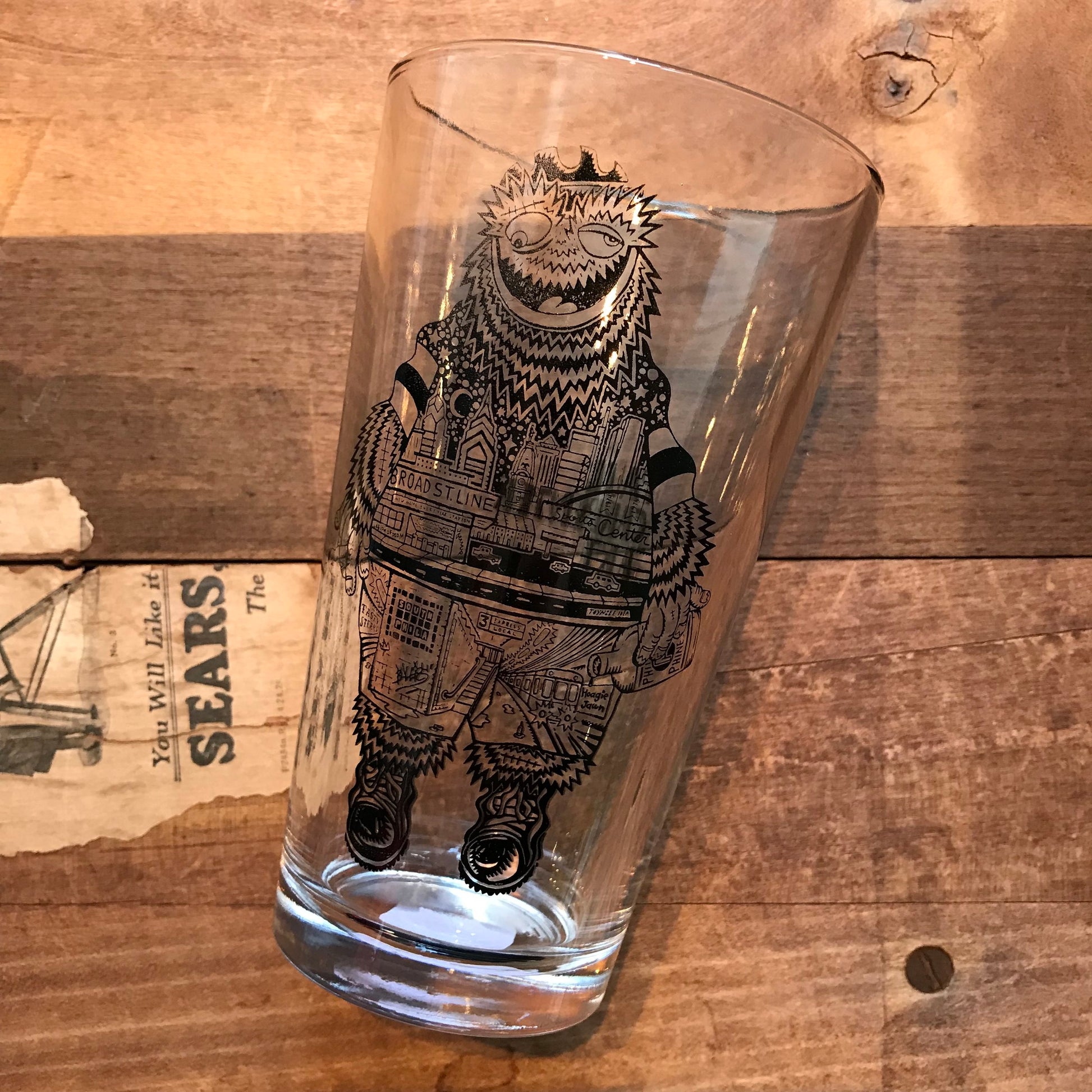A Paul Carpenter Philly Pint Glass with an intricate illustration of a mythical creature and the city skyline printed on it, placed on a wooden surface.