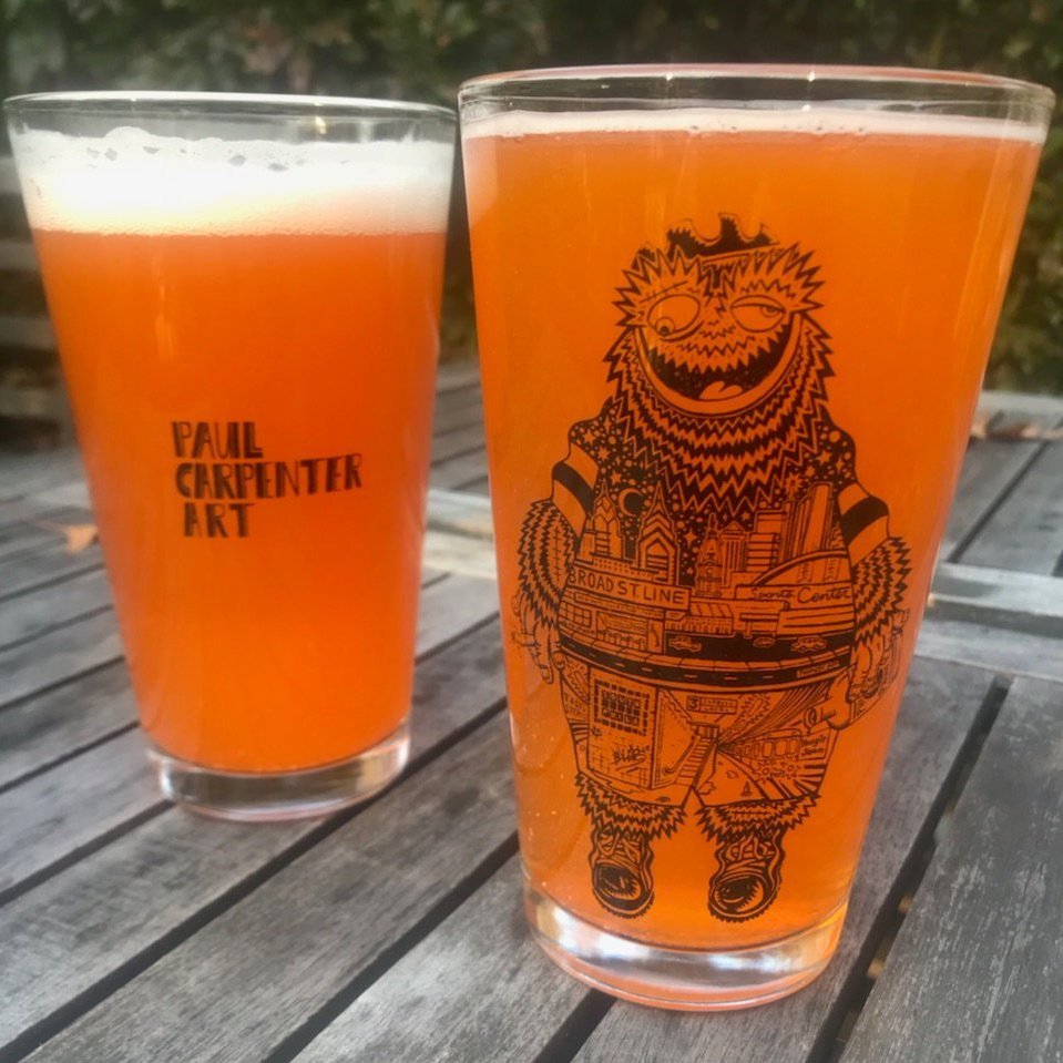 Two Philly Pint Glasses of amber-colored beer on a wooden table, with the nearer glass featuring an intricate black Philadelphia-themed skyline illustration by Paul Carpenter.