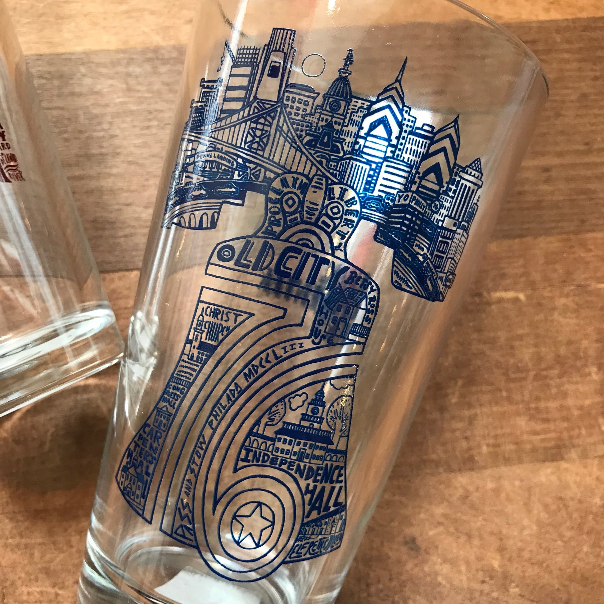 A Philly Pint Glass featuring blue illustrations and text that highlight landmarks and skyline associated with old city Philadelphia, Paul Carpenter-themed.