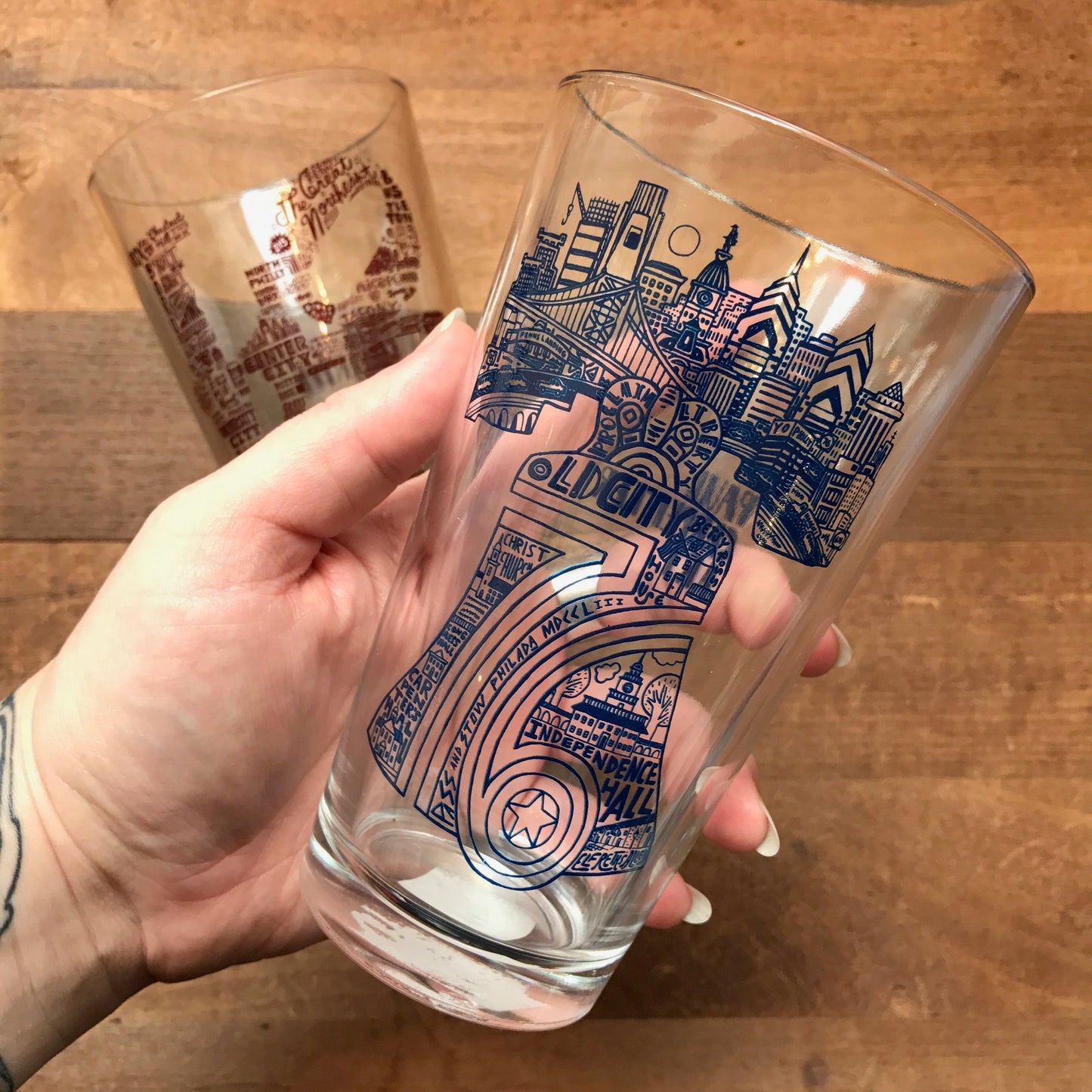 A hand holding a transparent Philly Pint Glass with intricate Philadelphia-themed skyline designs and text by Paul Carpenter.