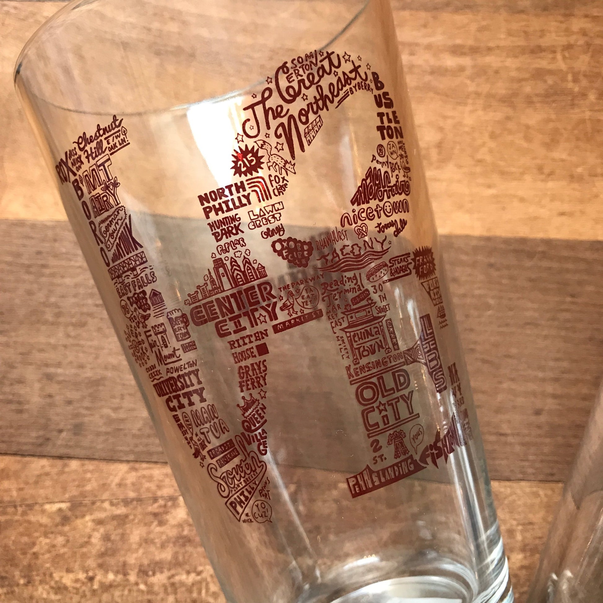 Clear Philly Pint Glasses with red text featuring various Philadelphia-themed neighborhood names and skyline landmarks by Paul Carpenter.
