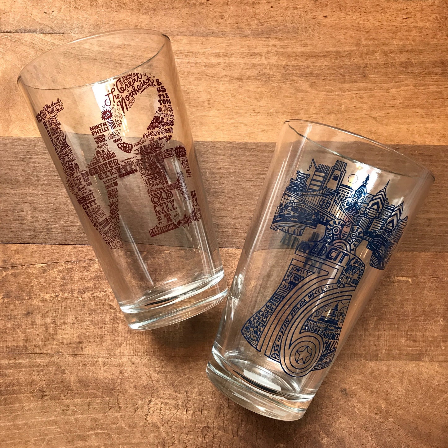 Two Philly Pint Glasses with Philadelphia-themed skyline illustrations on a wooden surface by Paul Carpenter.