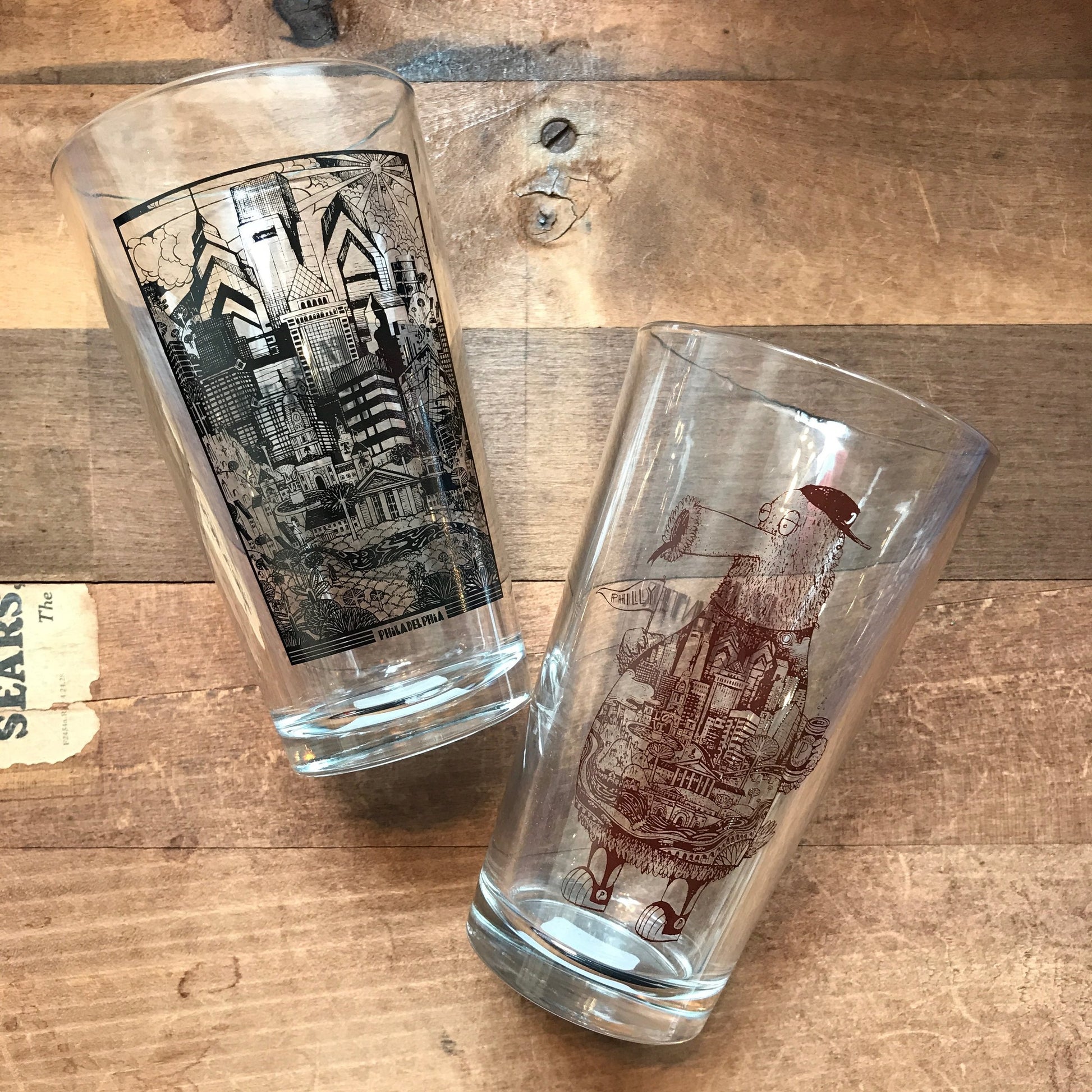 Two Paul Carpenter Philly Pint Glasses laying on a wooden surface, one upright with a Philadelphia-themed skyline design and the other tilted with a ship illustration.